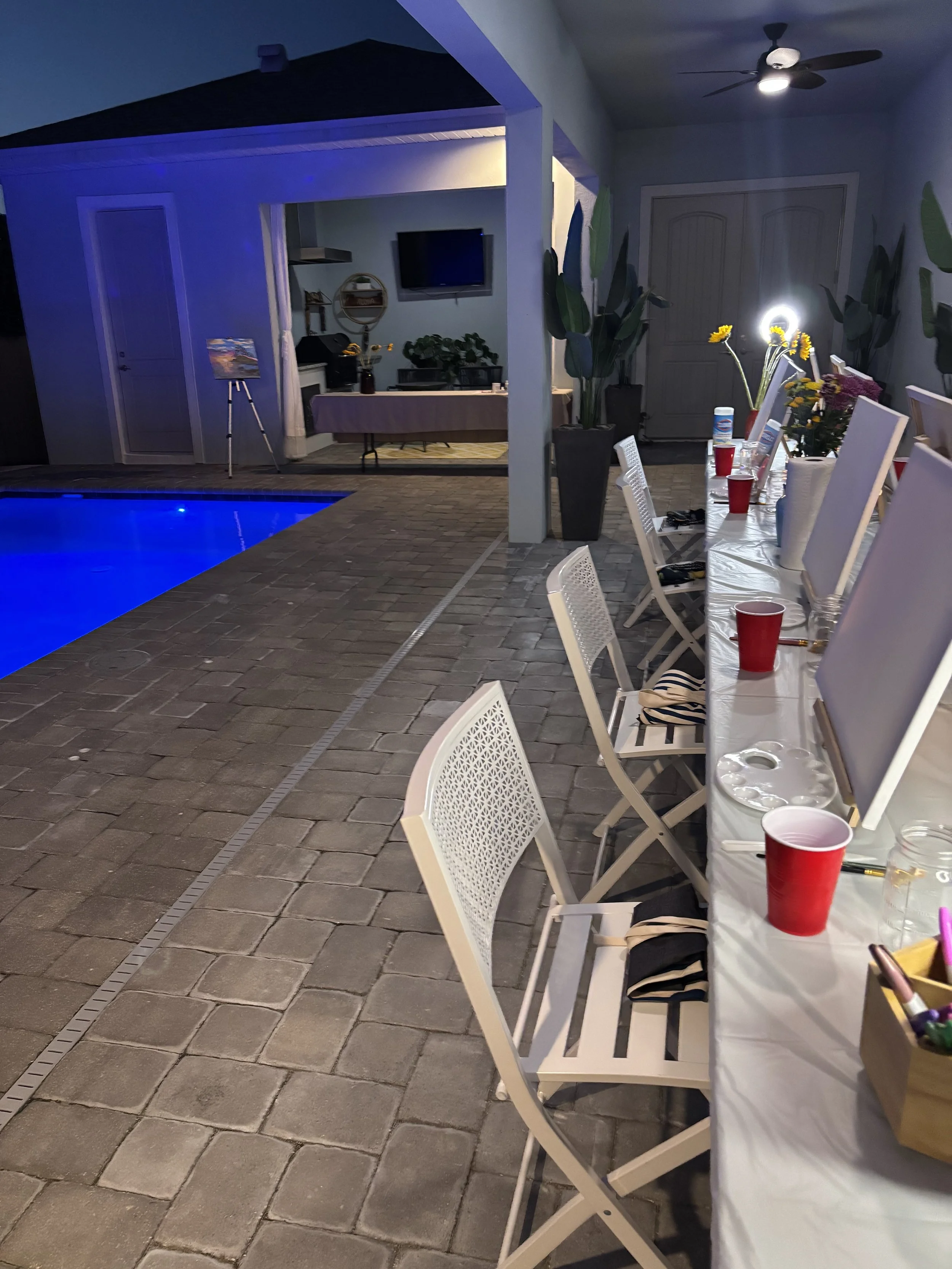 A nighttime outdoor party setup with a long table decorated with red cups, vases with yellow and purple flowers, and chairs. There are potted plants, a pool with blue lighting, and a double door in the background. The area is lit with ceiling lights and a wall-mounted light.