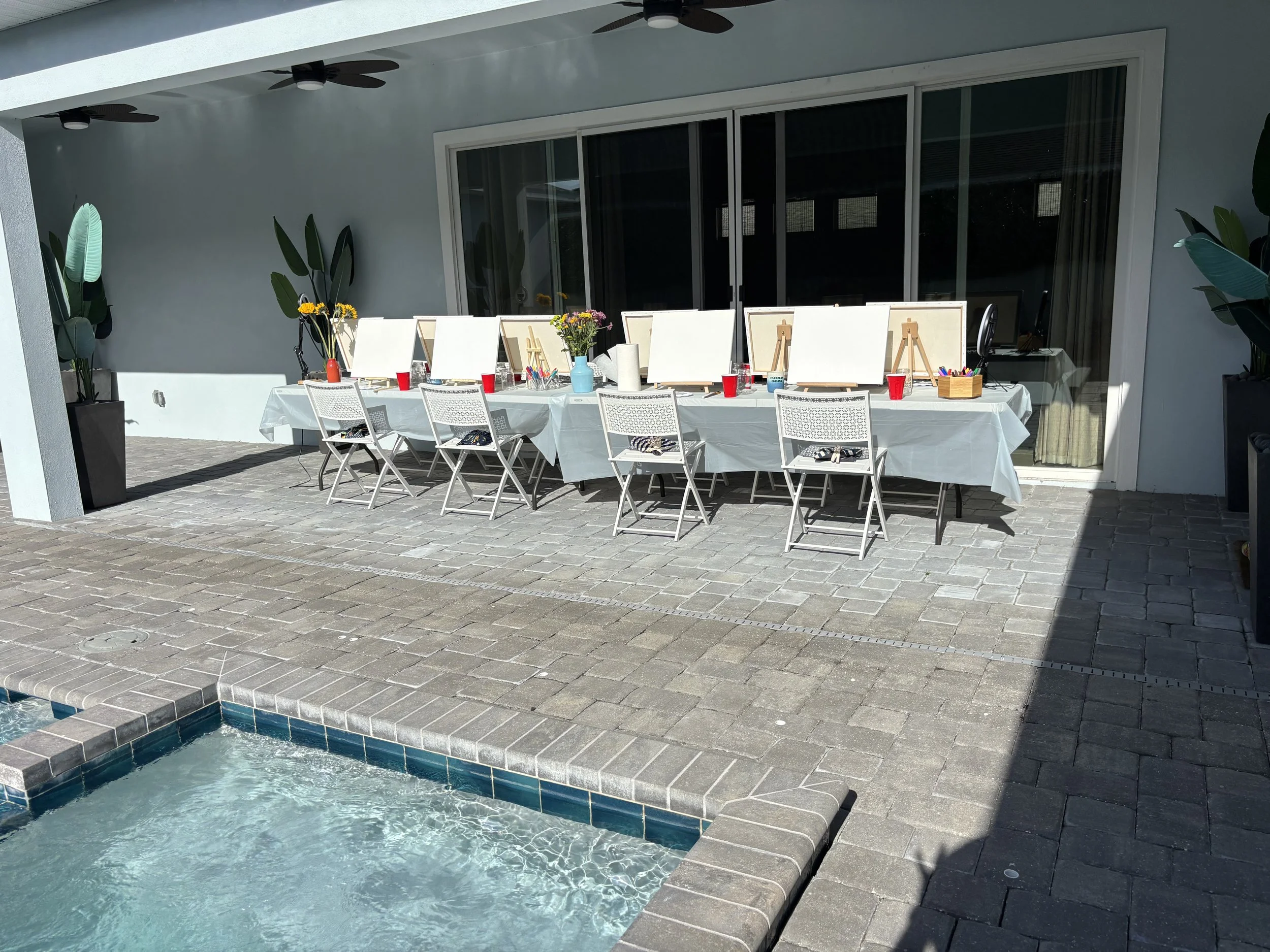 Outdoor patio setup with a long table covered with a white tablecloth, chairs, and painting supplies for a painting party, near a pool with water and brick paving.