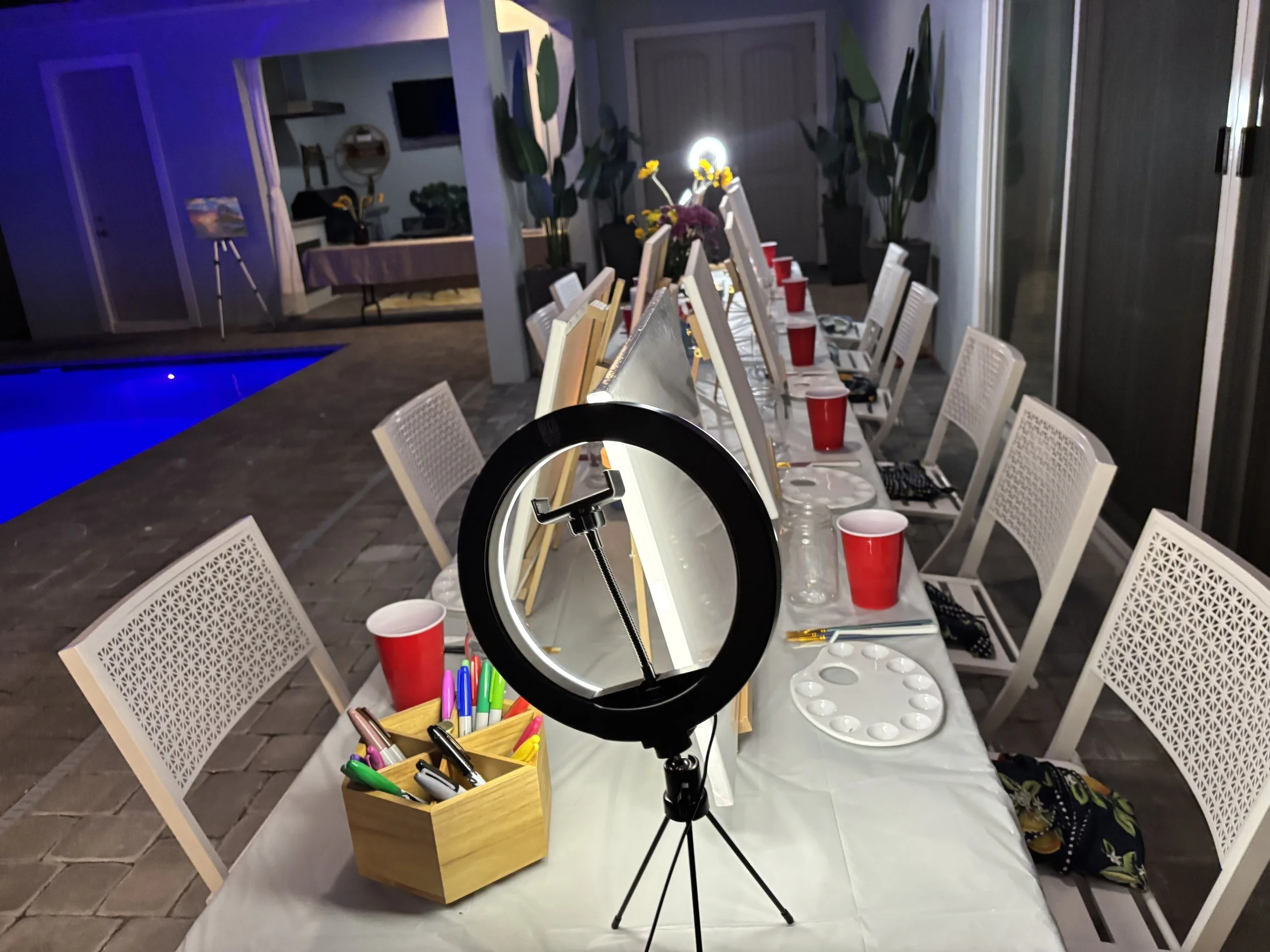 A long table set up for a painting party, with canvases, paint palettes, brushes, and cups, near a swimming pool at night.