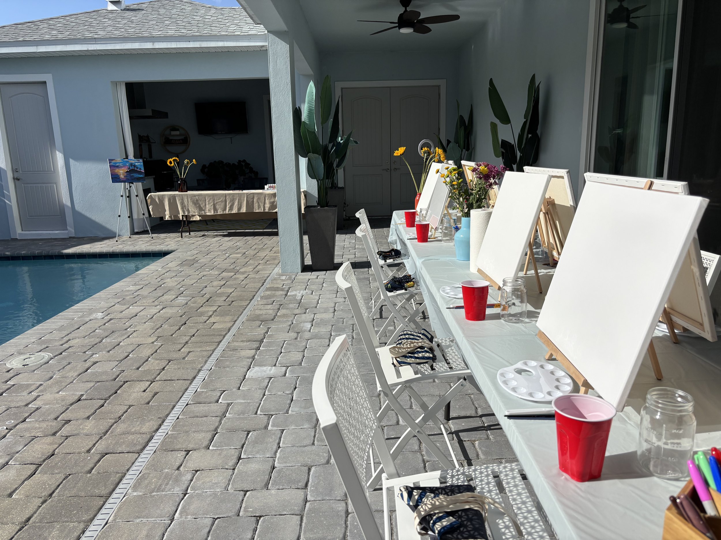 Outdoor patio with a pool on the left, and a long table on the right covered with white tablecloth, art supplies, red cups, vases, and flowers, with chairs lined up along the table. In the background, there are plants, a TV, and an easel with a colorful painting.