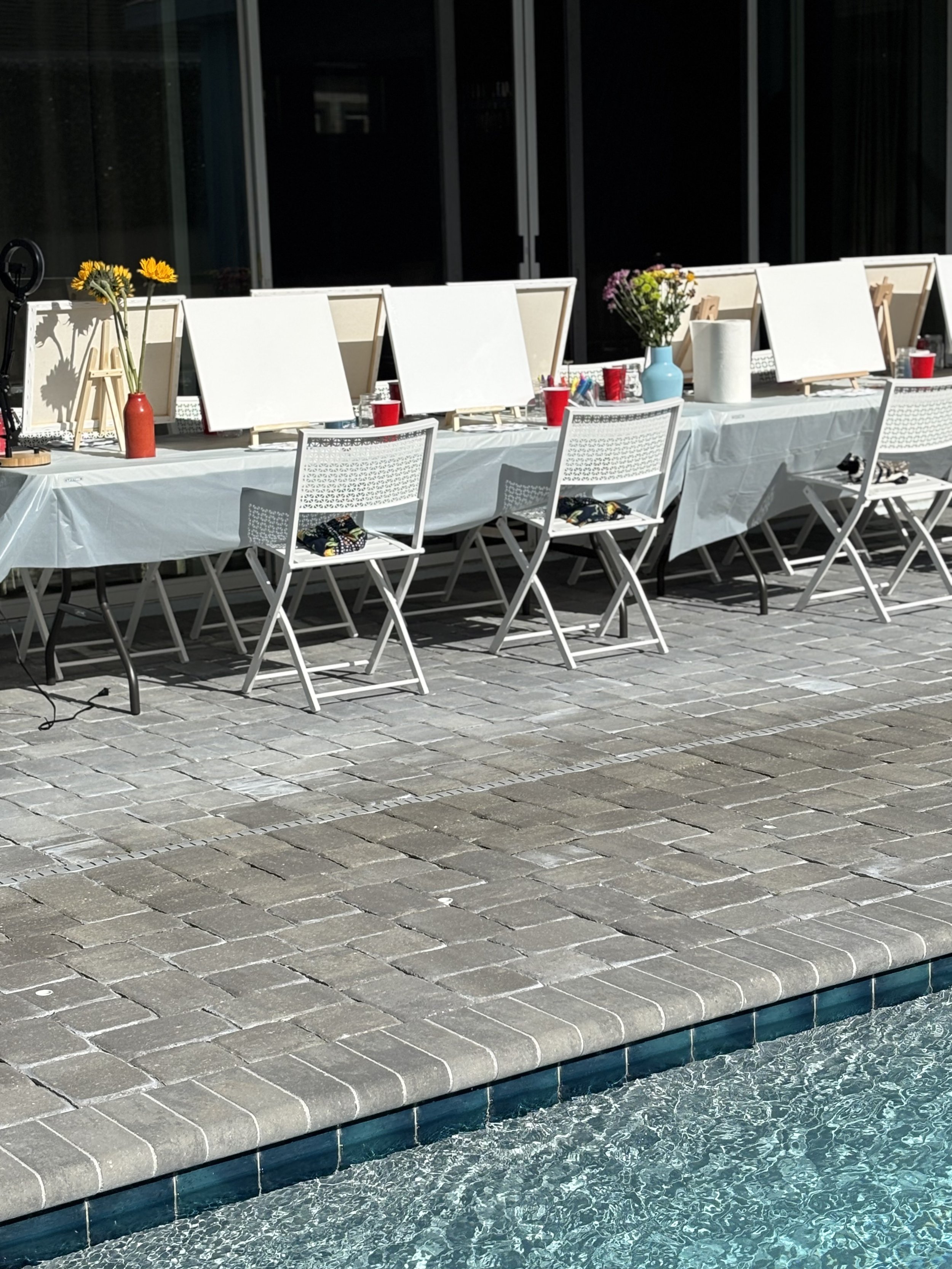 Tables set up for painting or art activity near a swimming pool, with blank canvases, easels, and supplies.