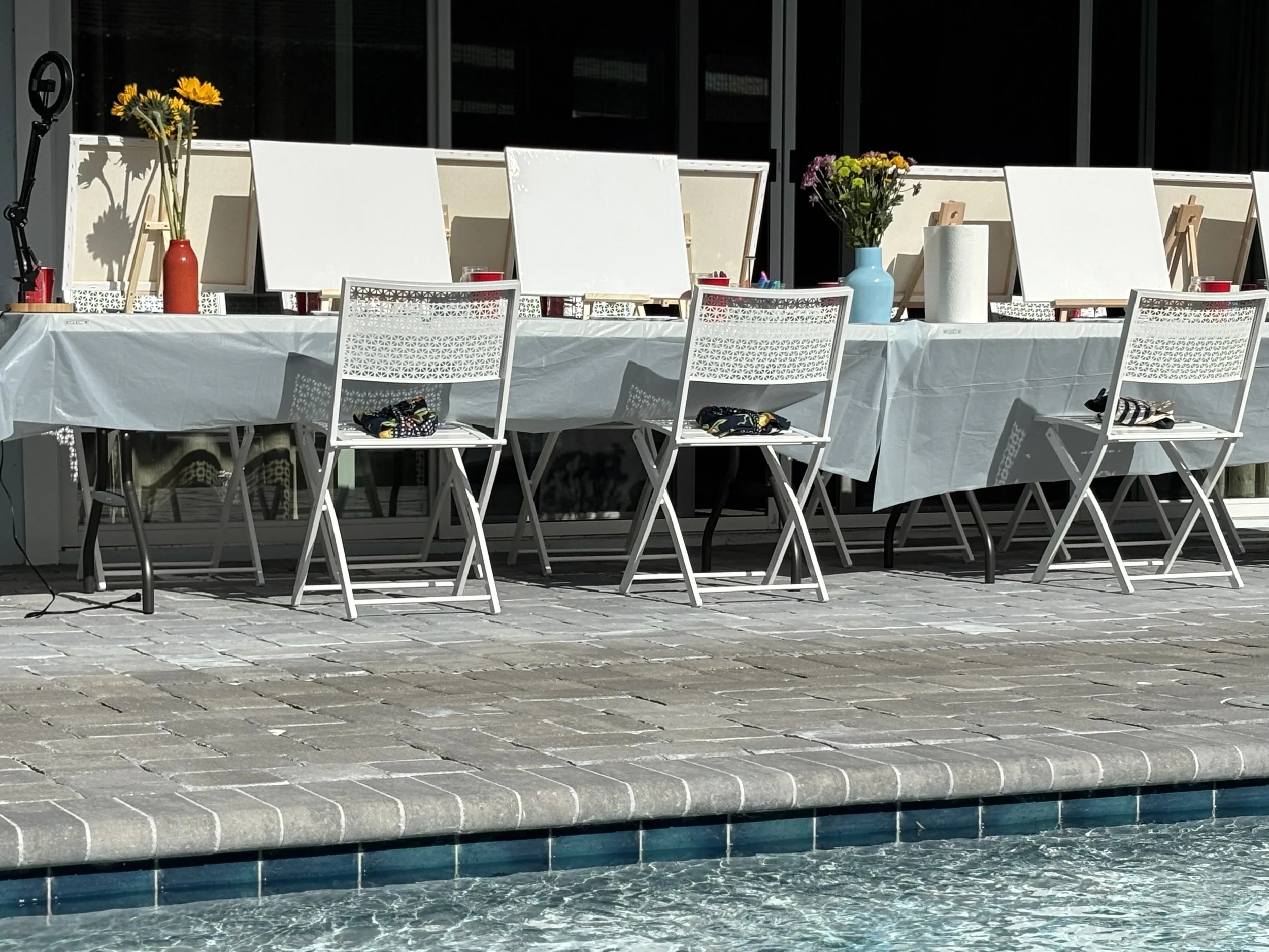 Empty outdoor table with white chairs and art supplies set up near a pool, with several colorful flower arrangements on the table.