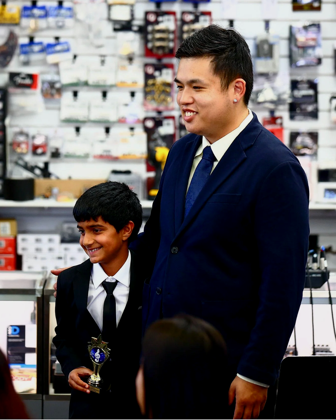 A young boy in a suit holding a trophy standing next to a man in a suit, in a store with various items and tools on shelves in the background.