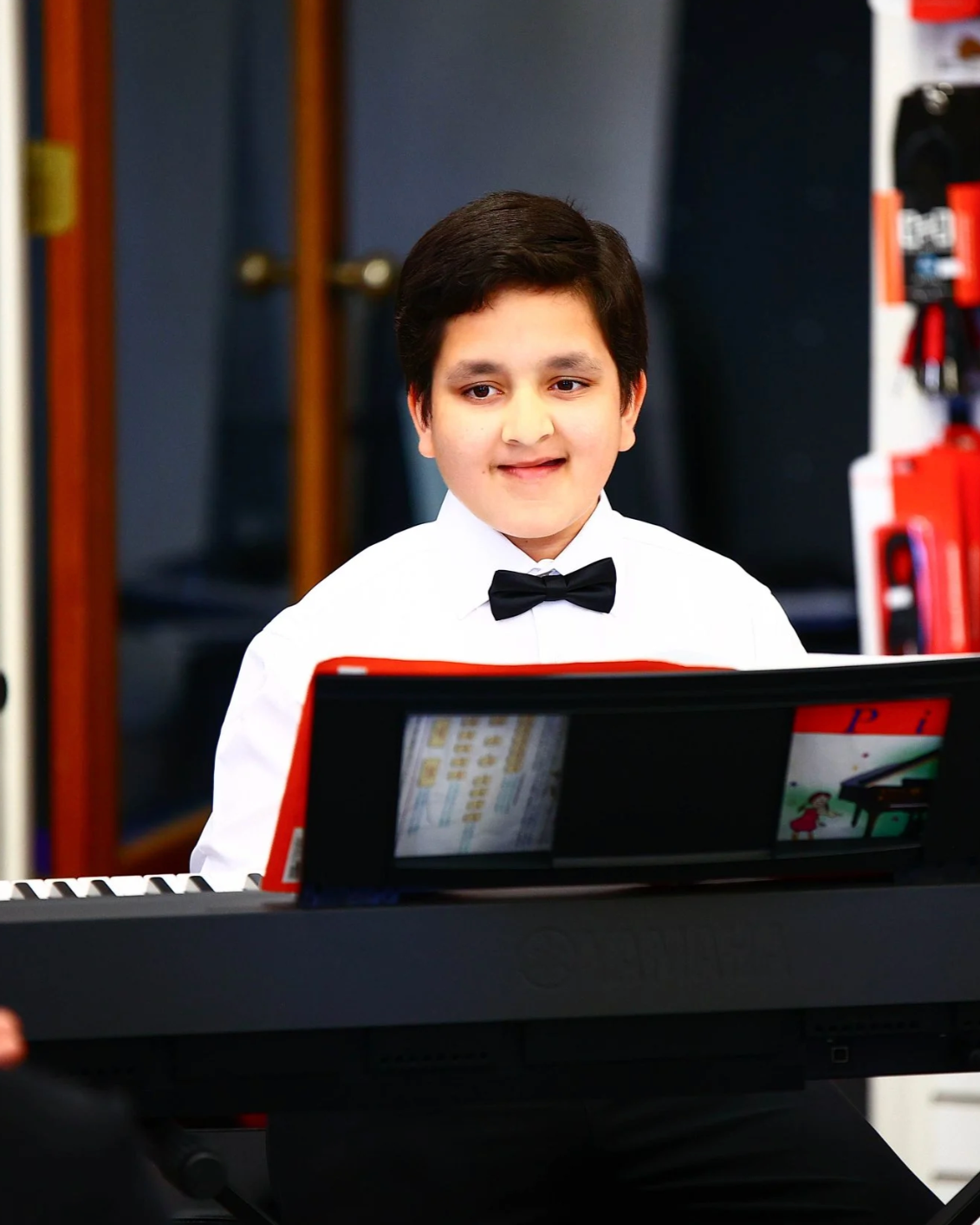 A young boy dressed in a white shirt and black bow tie playing a keyboard in a room, possibly during a performance or practice session.