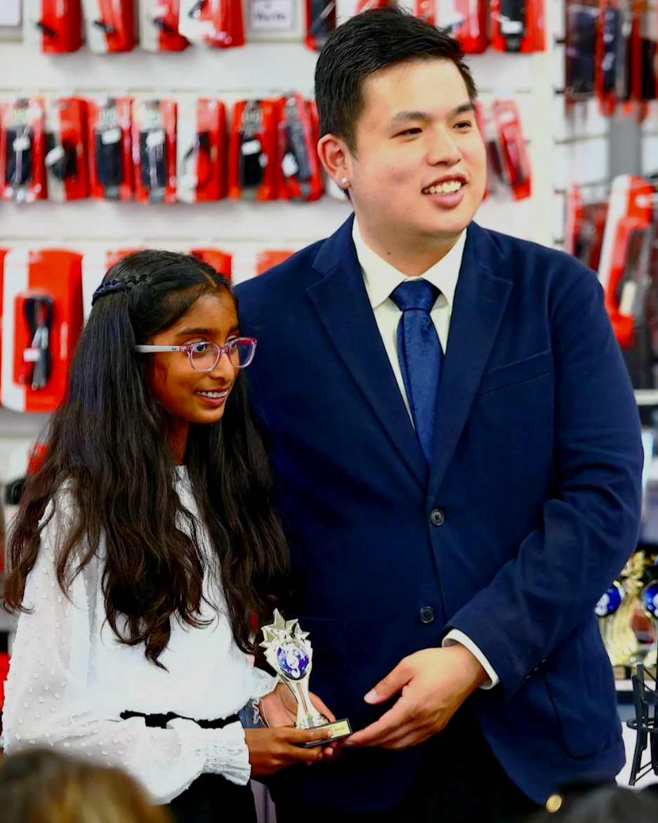 A young girl and a man in formal attire standing together in an electronics store, the girl holding a small trophy and smiling.