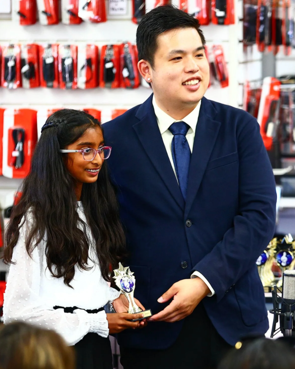 A young girl holding a trophy standing next to a man in a navy blue suit, in a store with red products on shelves.