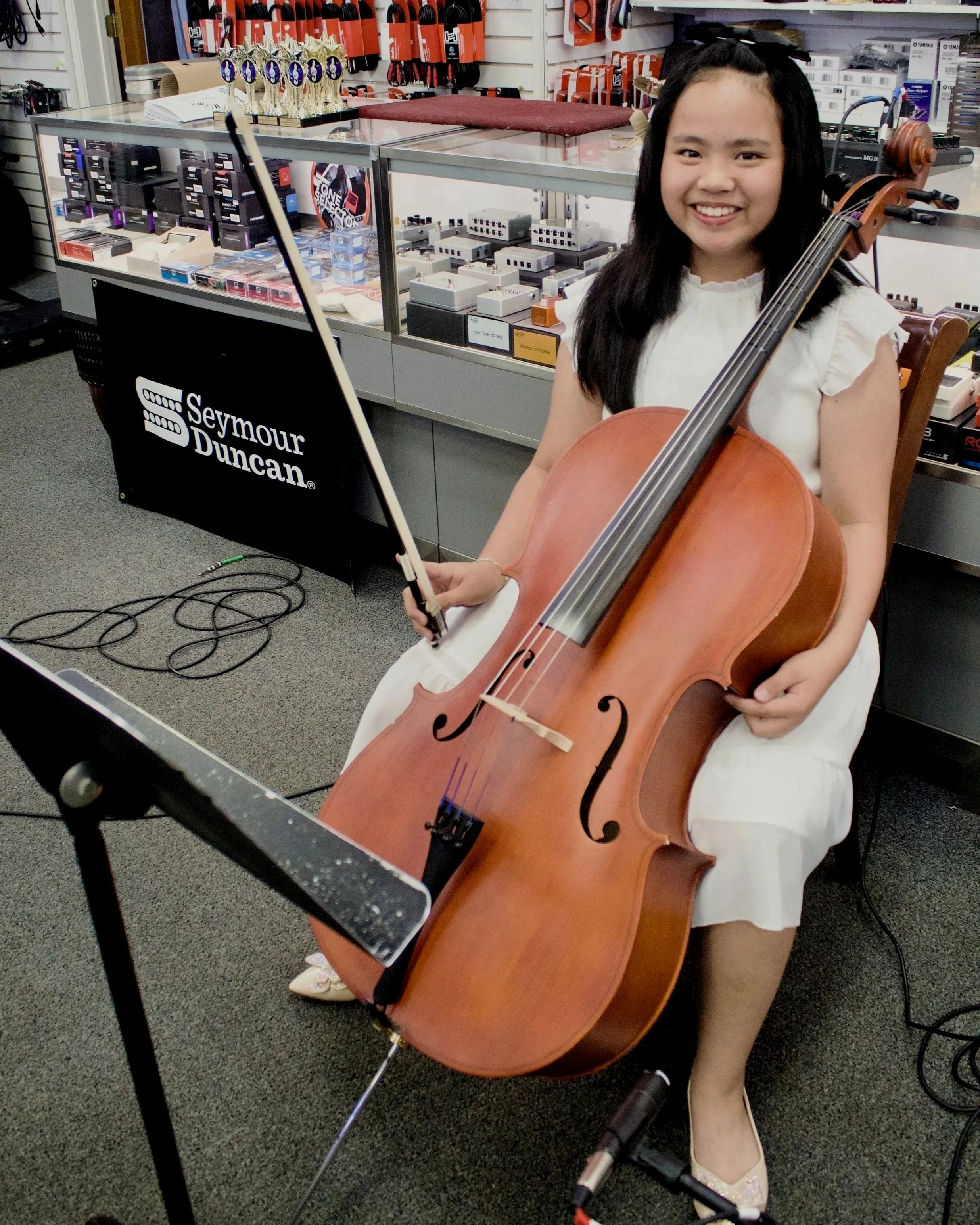 A young girl smiling while holding a cello and bow in front of a display case with musical equipment and medals in a music store.