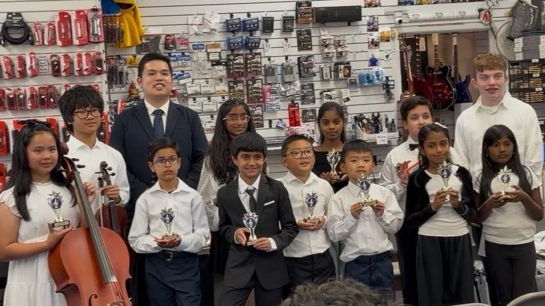 Group of children holding trophies, some wearing formal attire, in a music store with musical instruments and accessories on the walls.