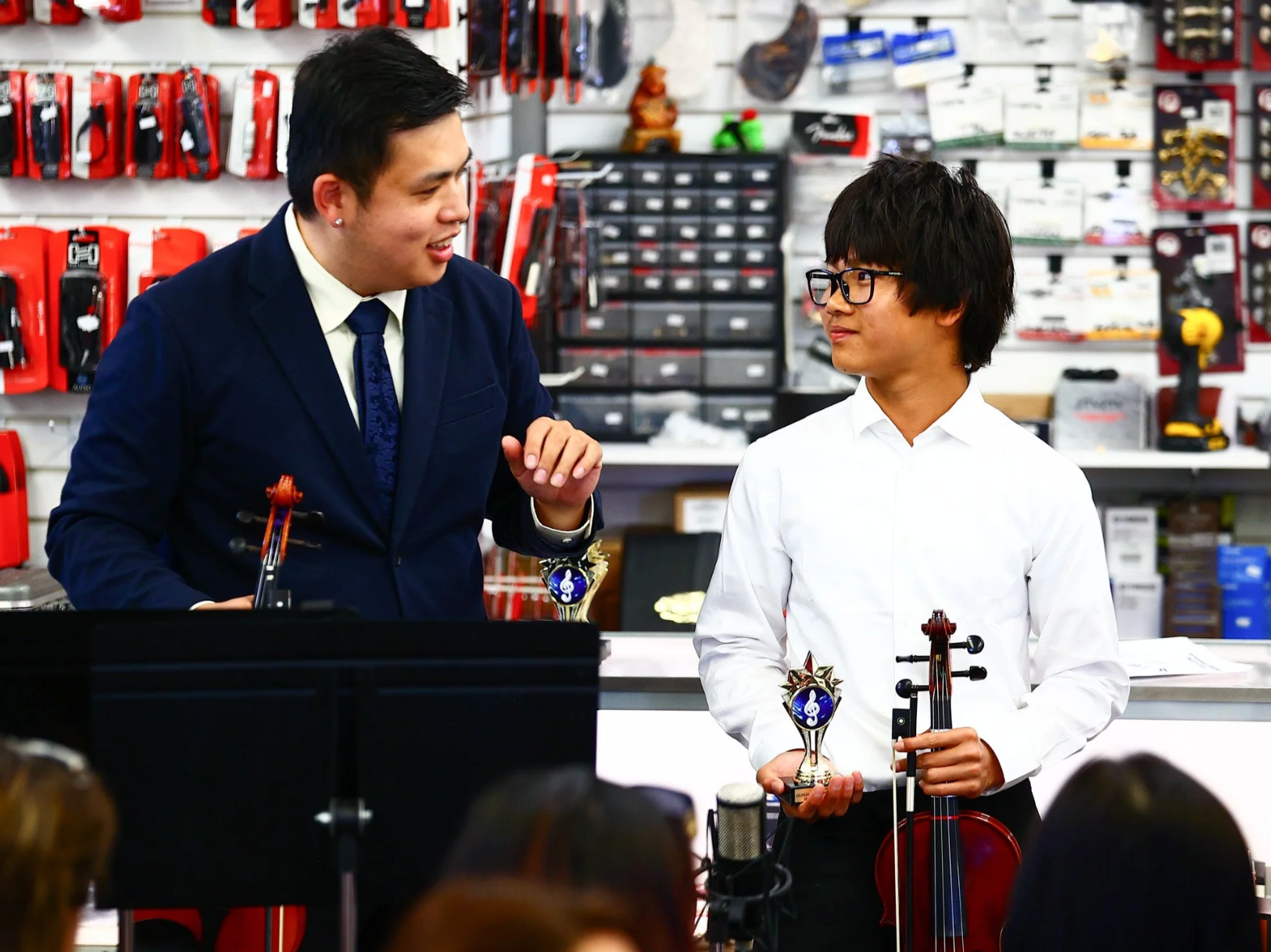 A man in a navy suit and a white shirt with a bowtie, and a young man in a white shirt and glasses, stand inside a store. The young man holds a trophy and a violin. They are engaged in conversation, with shelves of musical accessories visible in the background.