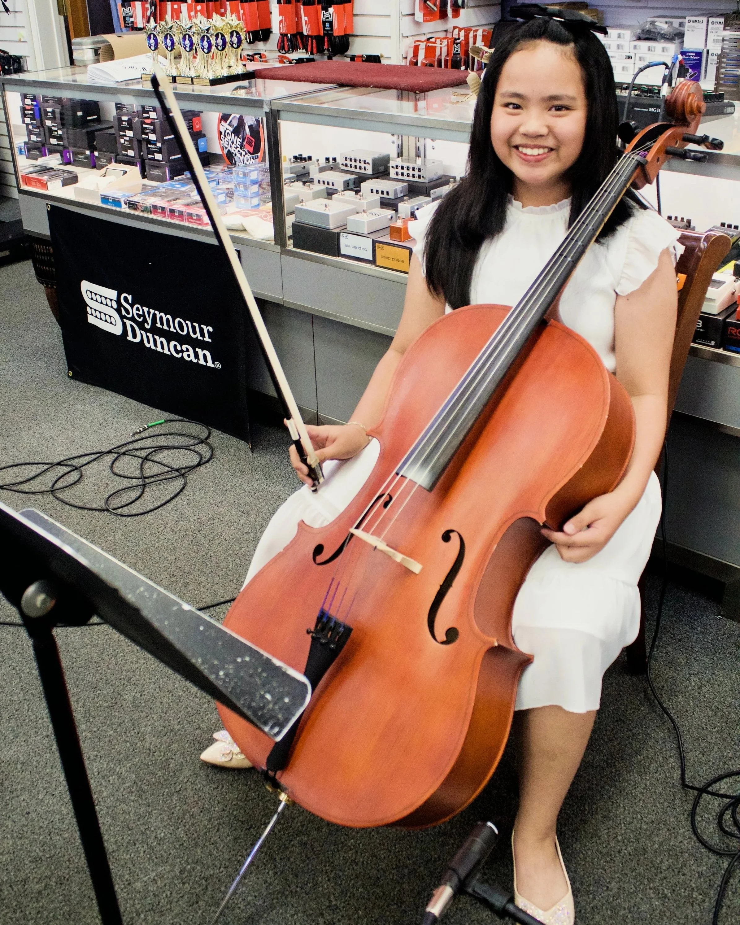 Young woman smiling and playing a cello in a music store, with shelves of musical accessories and trophies visible in the background.