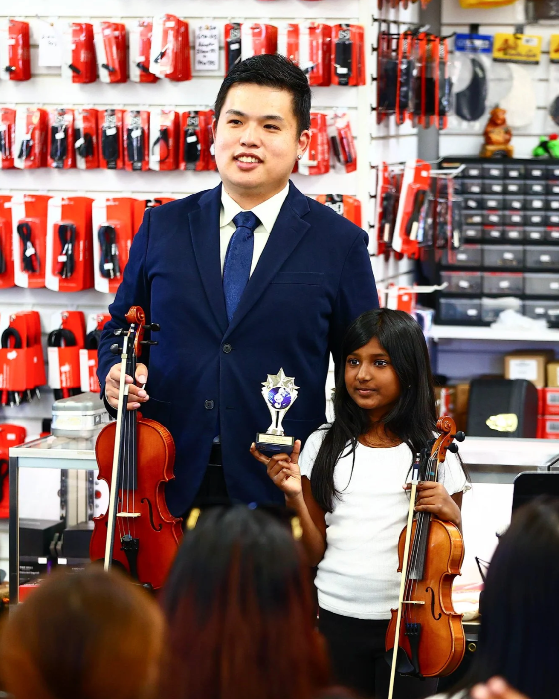 A young girl and a man posing in a music store, holding violins and a trophy, with red items on the wall behind them.
