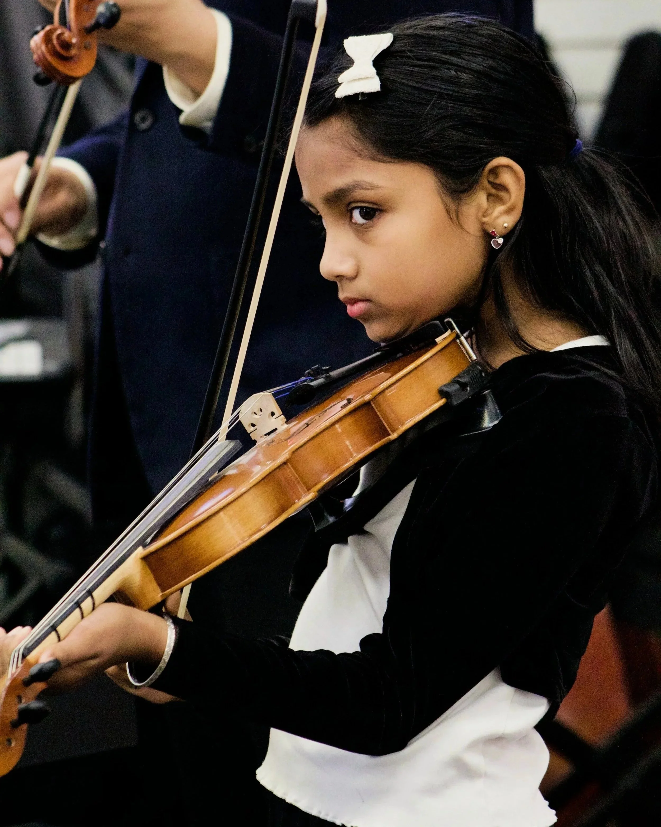 A young girl with dark hair, wearing earrings, playing the violin during a performance or practice session.
