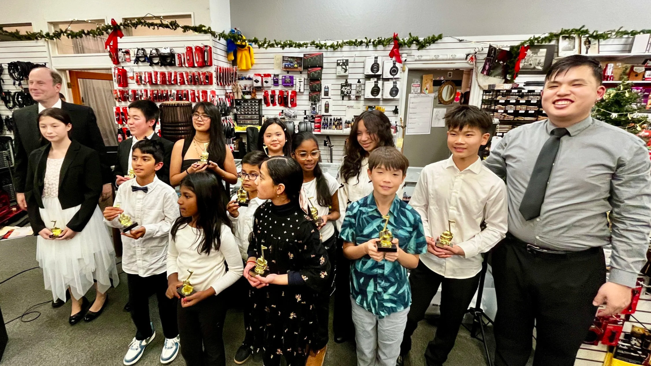 Group of children and adults at an award ceremony, holding trophies, in a store decorated with Christmas garlands.