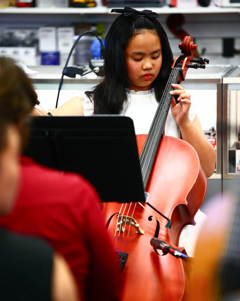 A young girl playing a cello during a musical performance in a store or classroom, with people and shelves in the background.