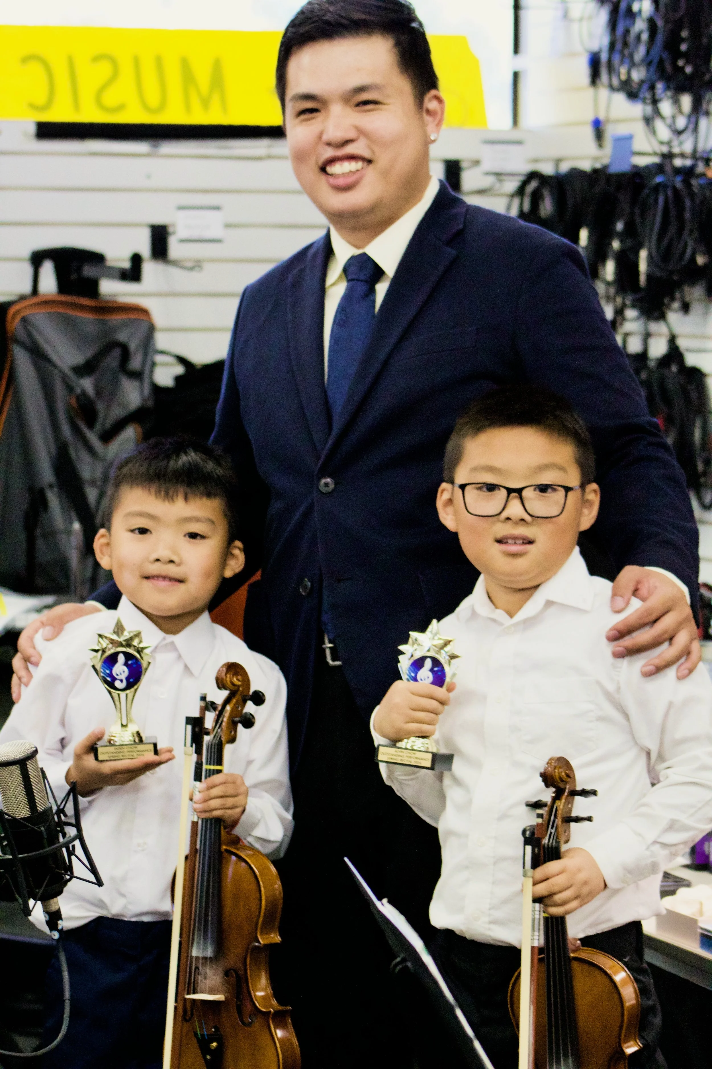 Two young boys wearing white shirts holding trophies, standing with a music teacher in a suit in a music store with musical instruments and equipment in the background.