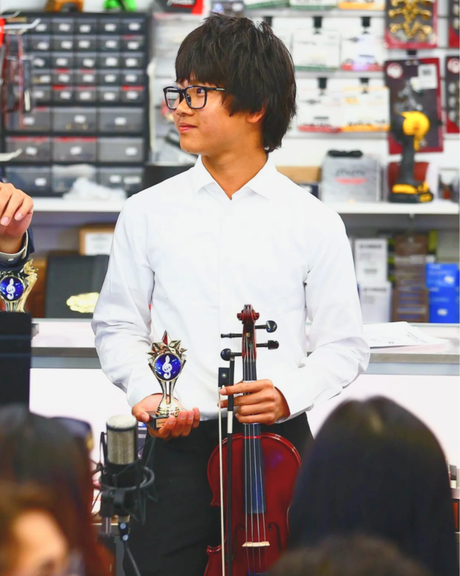 A young man with glasses in a white shirt holding a music award and a violin.