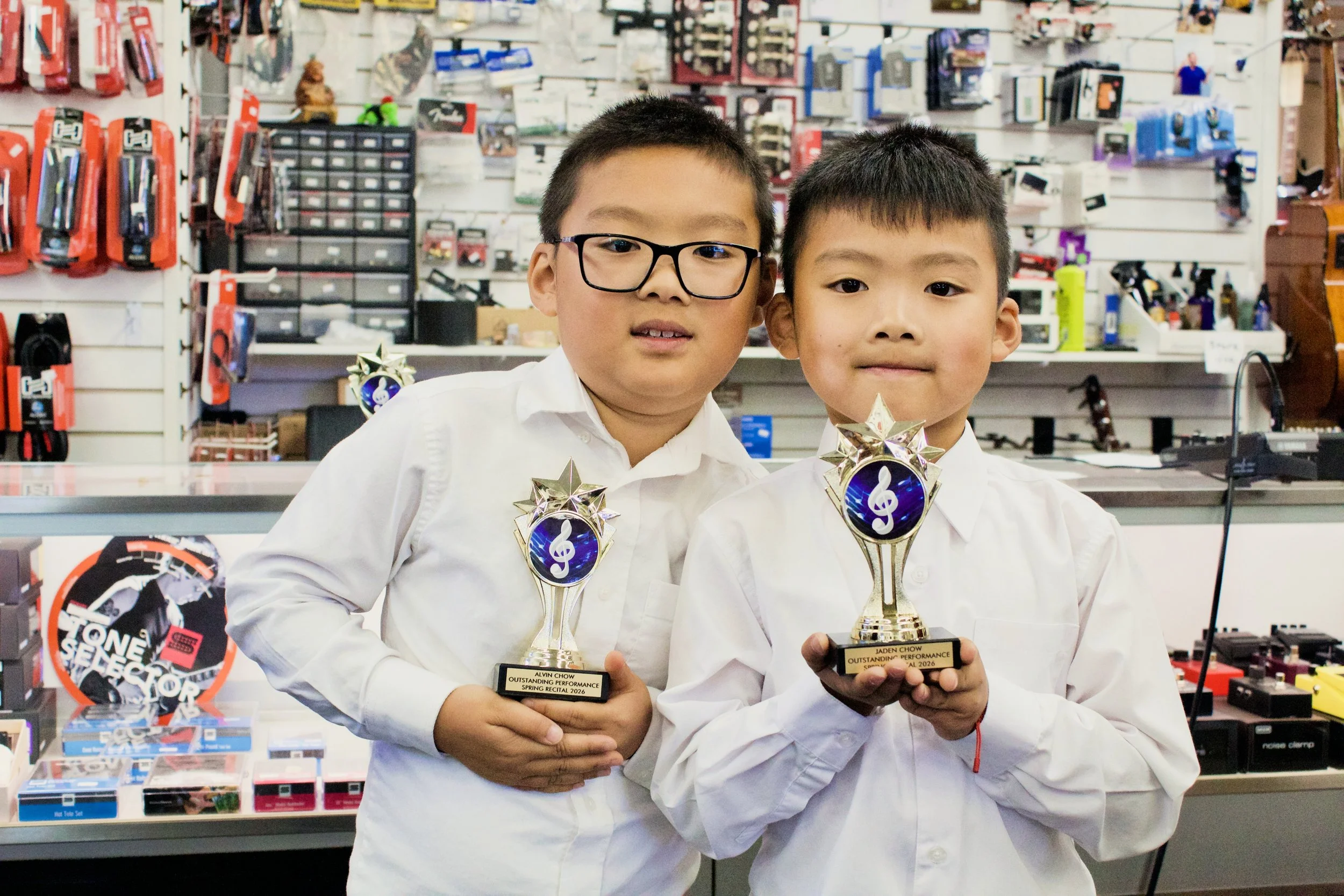 Two young boys with short black hair and glasses holding trophies in a store or music shop. The trophies have musical notes and stars on top, with labels reading their names and achievement details. They are dressed in white shirts and seem proud of their accomplishments.