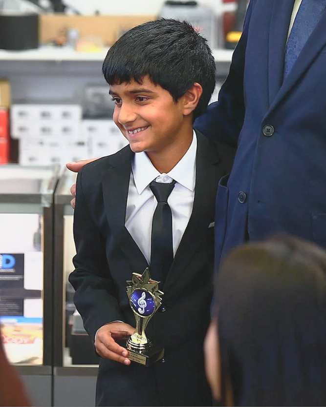 A young boy in a suit holding a trophy with a treble clef symbol, standing in an indoor setting, smiling, with a teacher next to him.