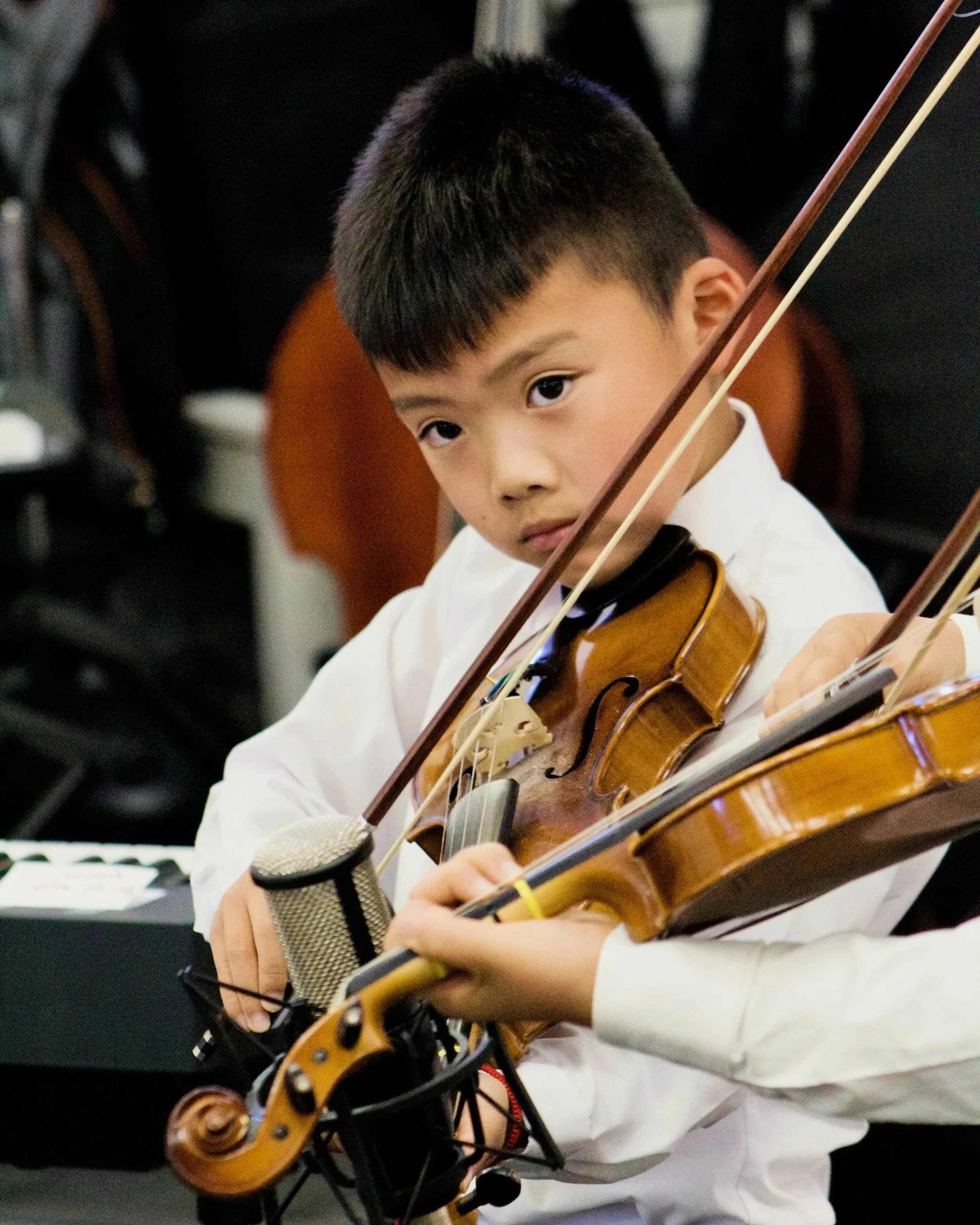 A young boy with short black hair is playing the violin, with a microphone nearby. He appears focused, and his face is partially turned, showing his right eye and part of his face. He is wearing a white shirt, and the background is dark with some indistinct objects.