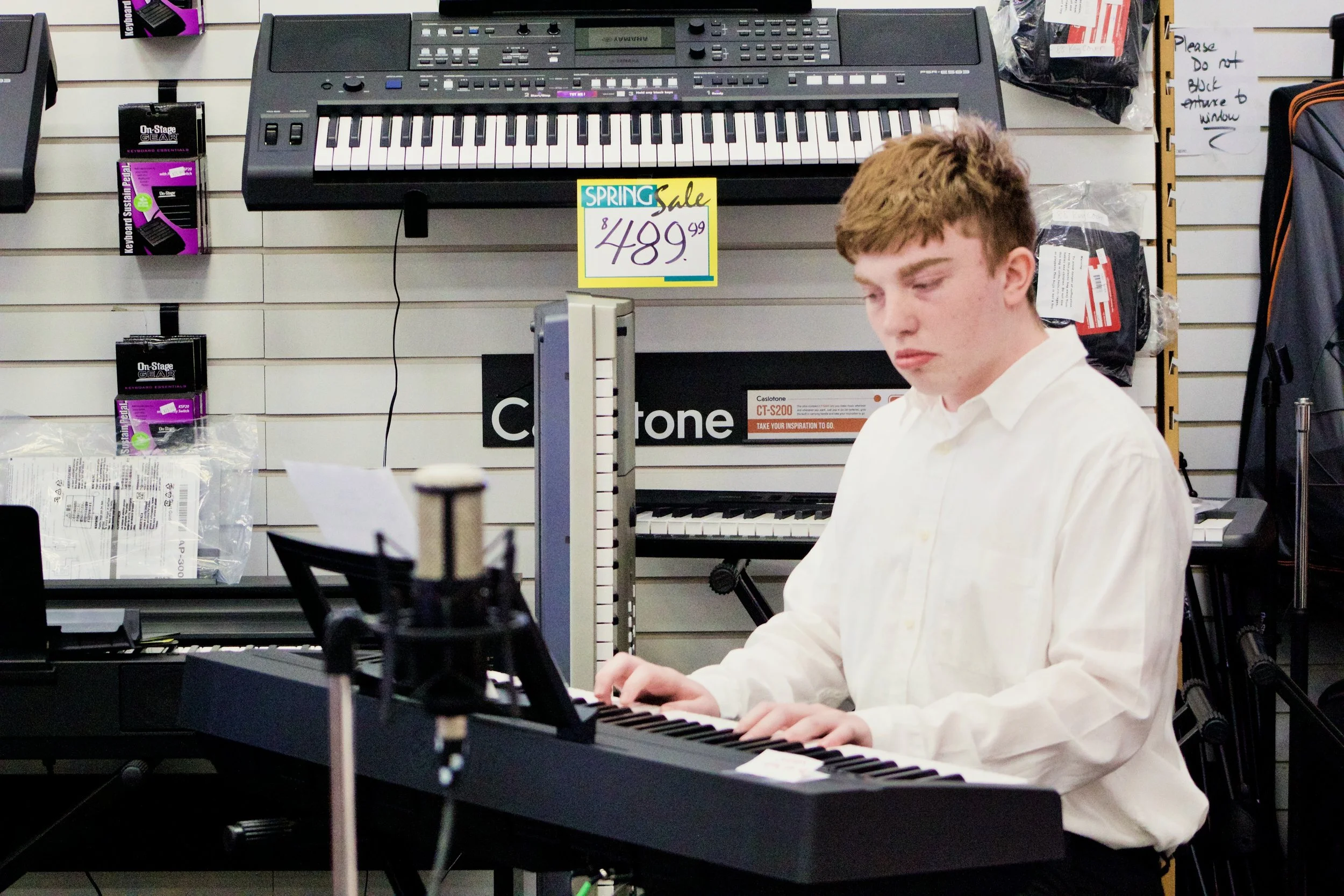 A young man with light skin and short, light brown hair wearing a white dress shirt playing a keyboard in a music store. The background features wall display racks with retail products, including a sign showing a spring sale price of $489.99 for the keyboard.