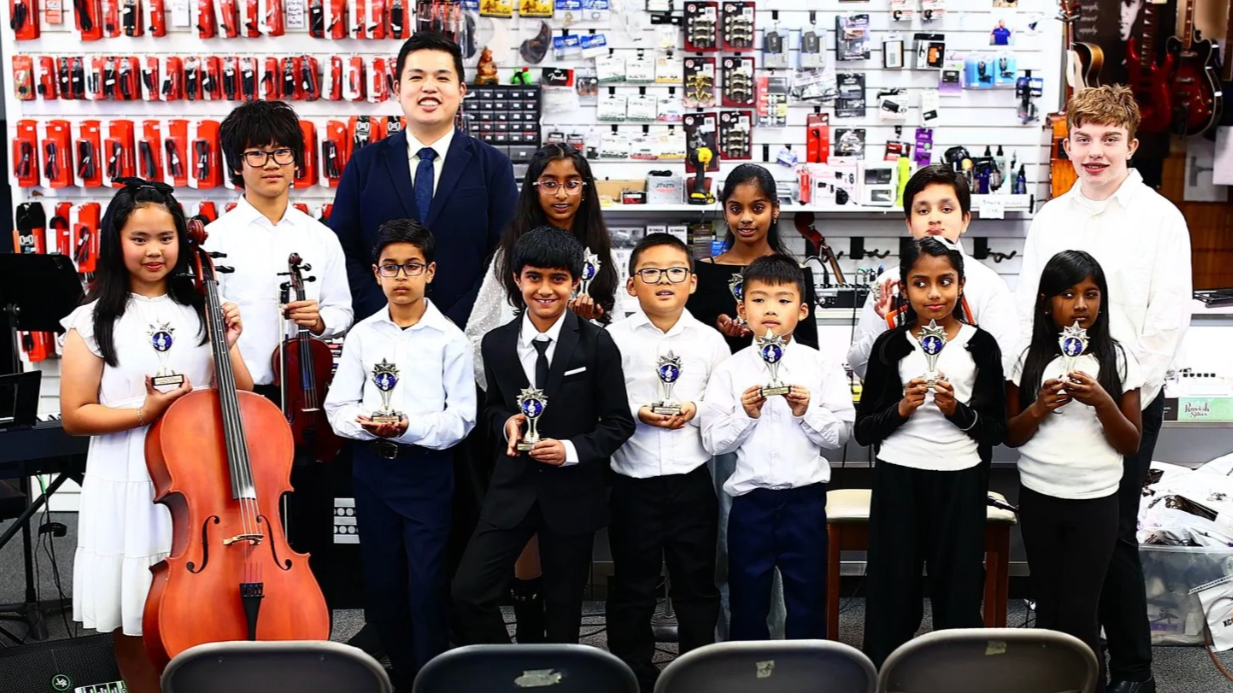 Group of young children in formal attire with a music teacher, holding trophies and musical instruments in a music store.