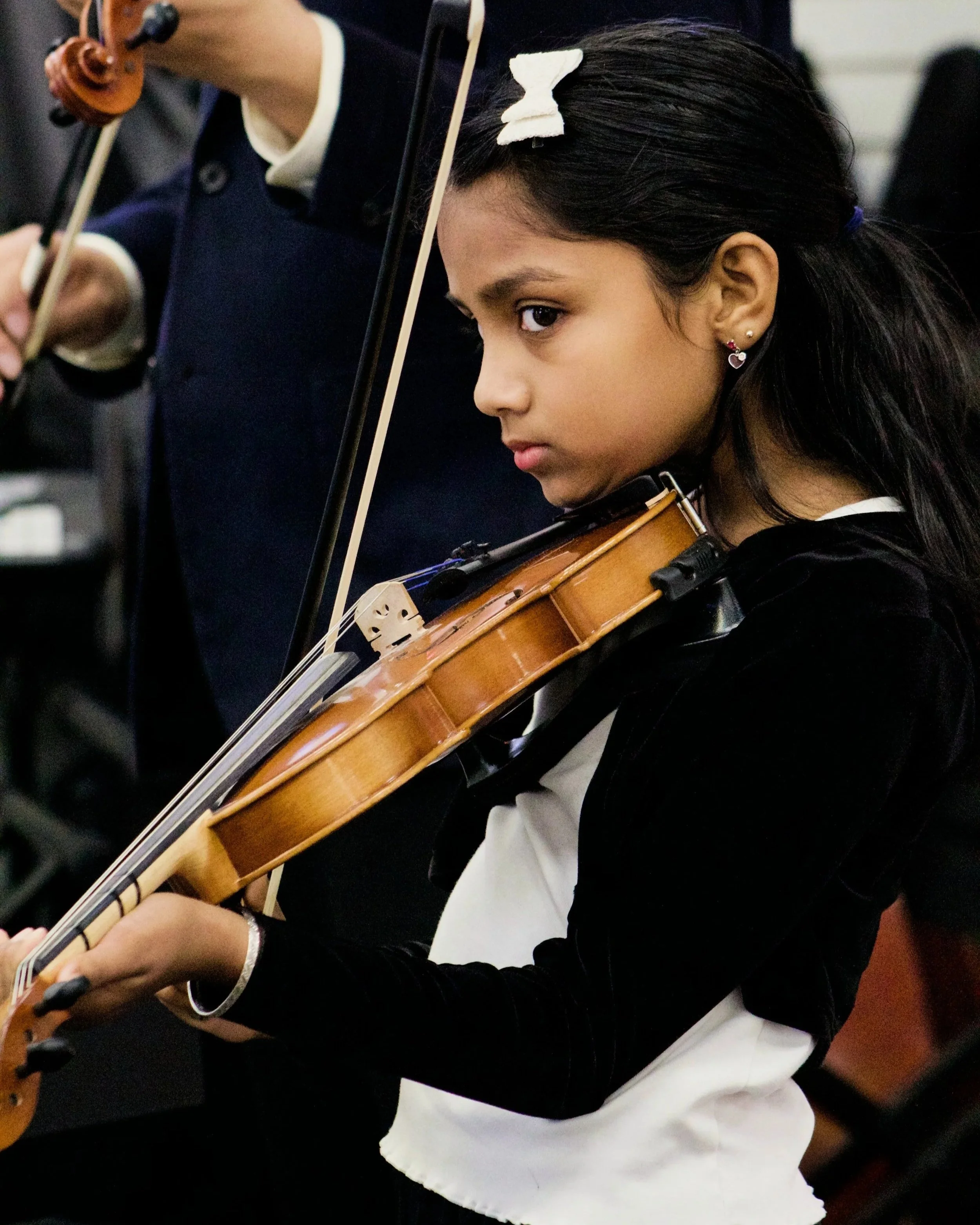 A young girl with dark hair, wearing a white bow, playing the violin, with a person in the background also playing an instrument.