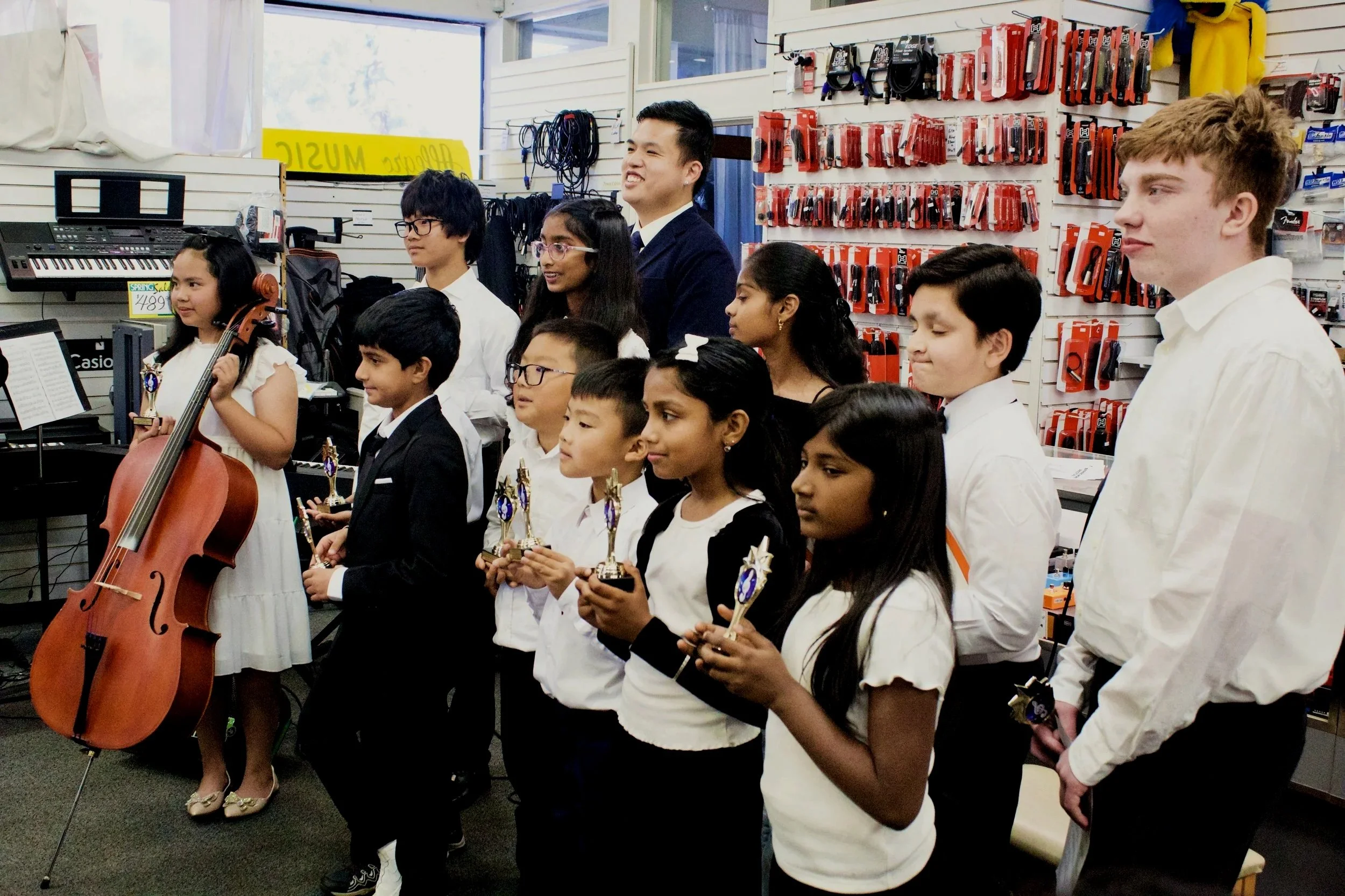 A diverse group of children and adults standing in a music store, holding trophies, with musical instruments and accessories on the walls behind them.