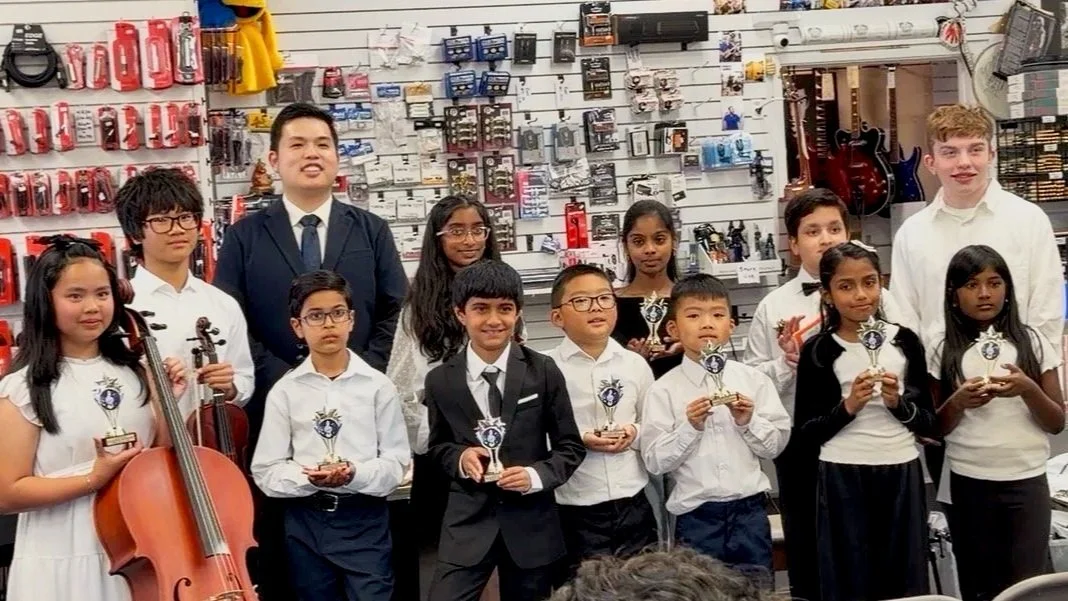 Group of children and two adults in musical attire holding trophies and musical instruments, standing in a music store or classroom with wall displays of guitar accessories and musical equipment.