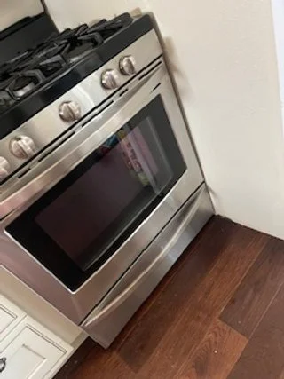 A stainless steel gas stove with oven in a kitchen corner, next to a white cabinet, on dark wood flooring.