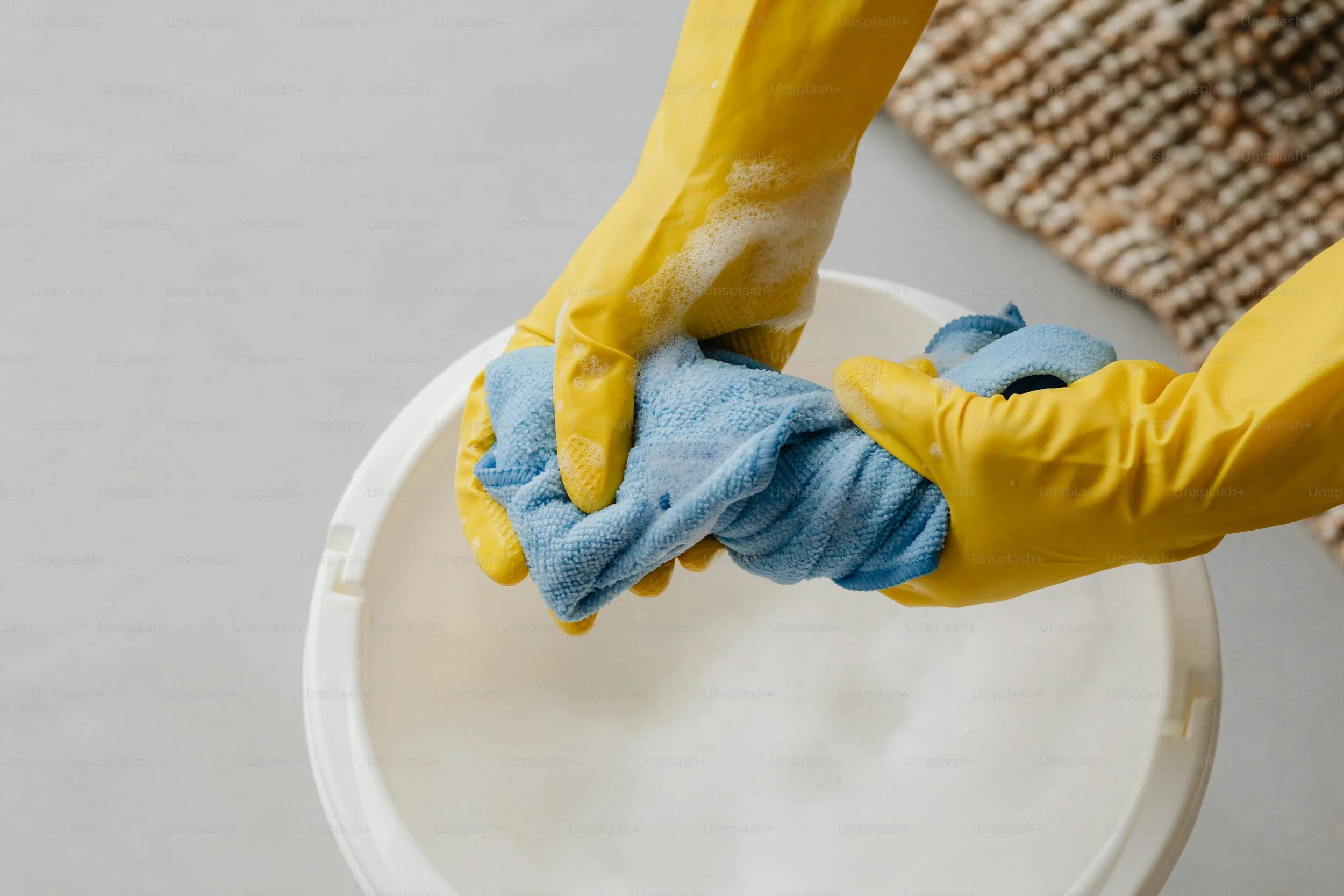 Person wearing yellow rubber gloves cleaning a white bucket with a blue cloth.