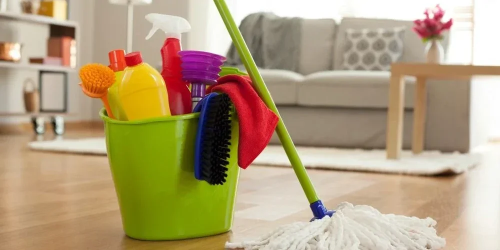 Cleaning supplies in a green bucket on a wooden floor next to a mop with a white mop head, in a living room with a sofa and a coffee table.