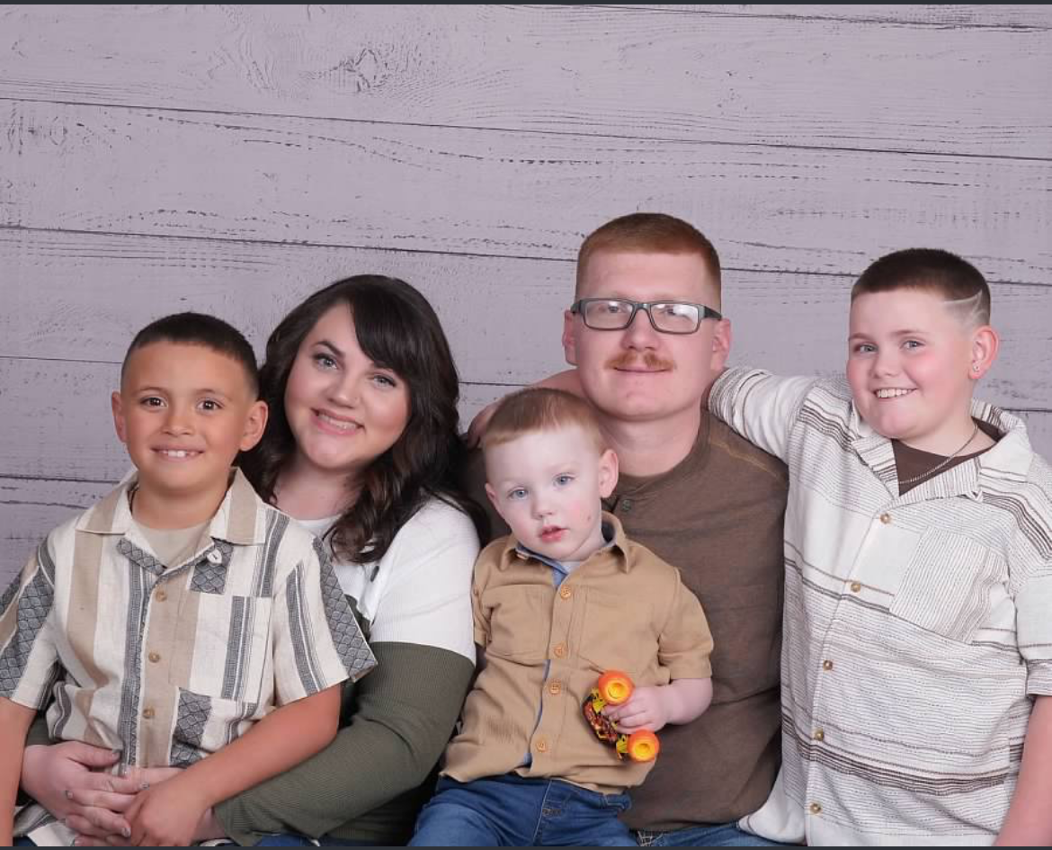 Family of five posing for a photo in front of a light wood backdrop, including two boys, a woman, a man, and a young child holding a toy.