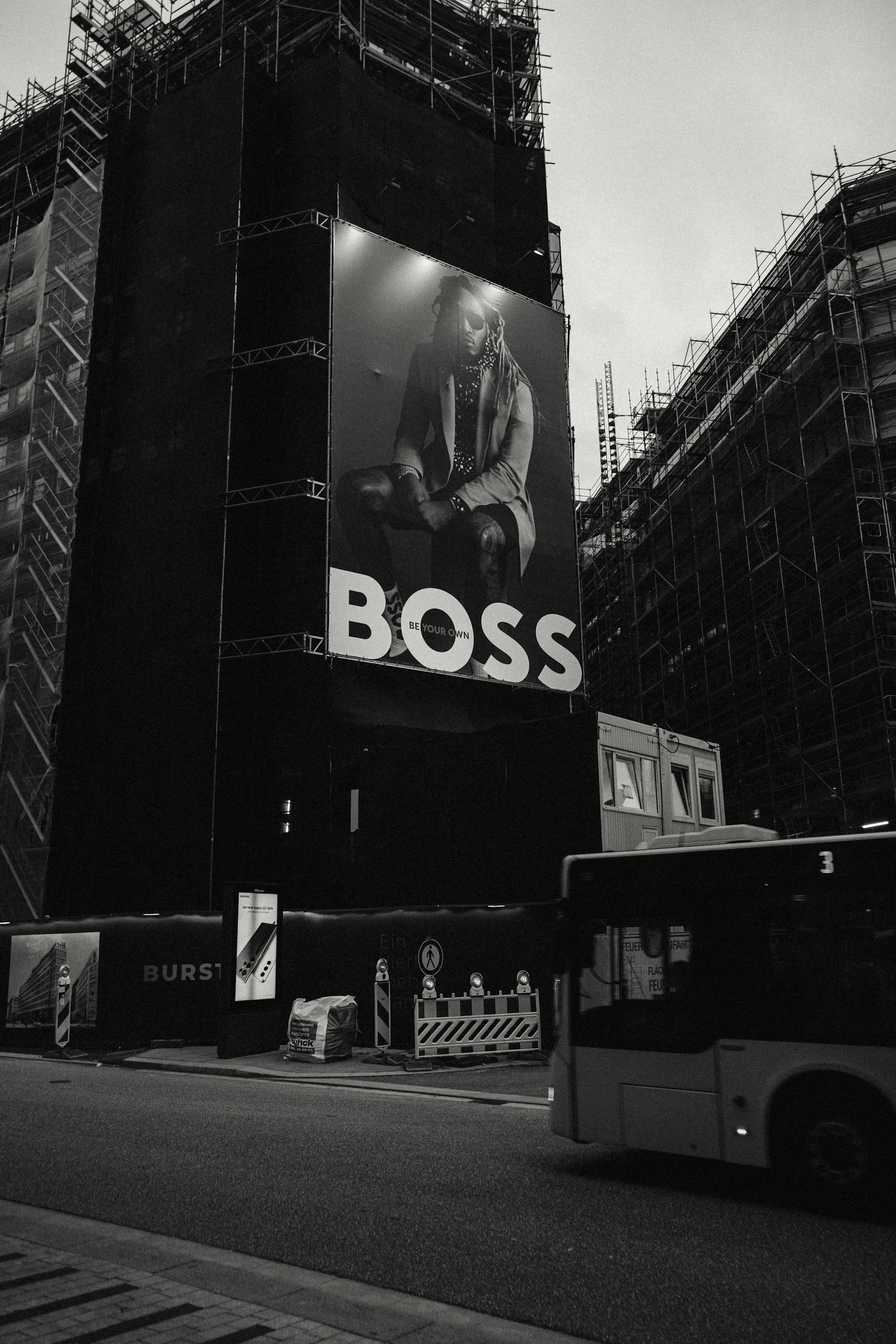 A black and white photo of a large billboard on a building under construction, featuring a model with dreadlocks, sunglasses, and fashionable clothing, with the word "BOSS" in bold letters at the bottom.
