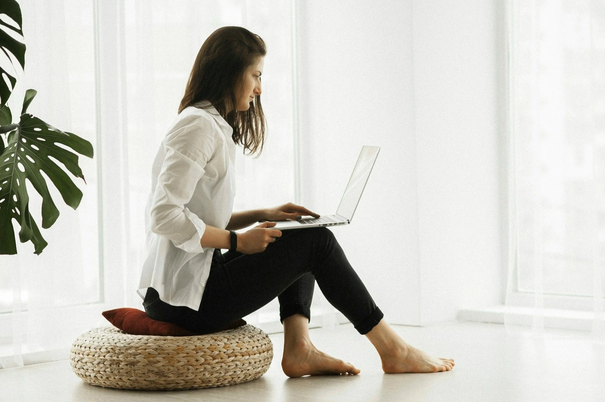 A woman with brown hair sitting on a woven pouf, working on a laptop in a bright, minimalist room with white walls and large windows, with green plant leaves in the background.