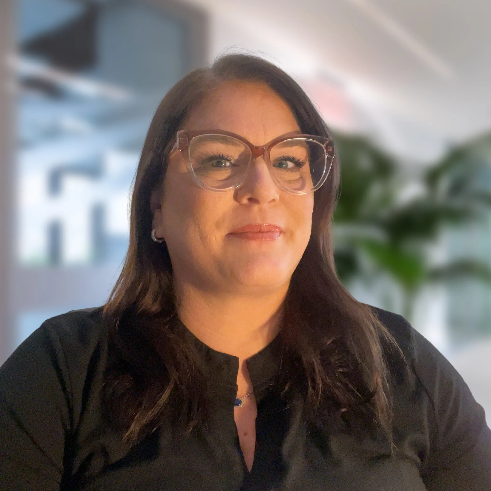 A woman with long dark hair, wearing glasses, a black top, and a necklace, smiling in a well-lit indoor setting with blurred greenery in the background.