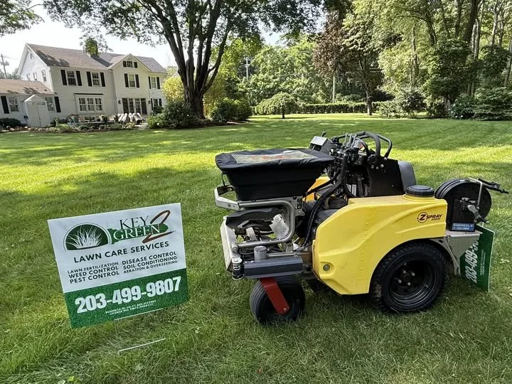 A yellow and black Z-Spray lawn treatment sprayer on a grassy lawn, with a white sign promoting Key Green Lawn Care Services nearby, and trees and residential houses in the background.