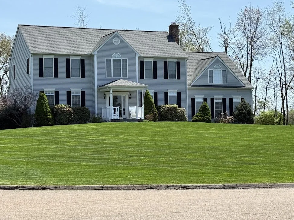 Large two-story house with light blue siding, black shutters, a small front porch, and a well-kept green lawn.