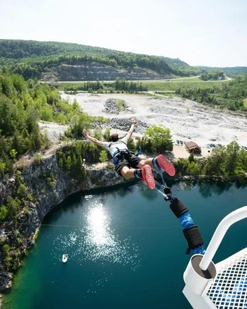 Person mid-air jumping from a quarry ledge into a body of water, with treeline and hills in the background.