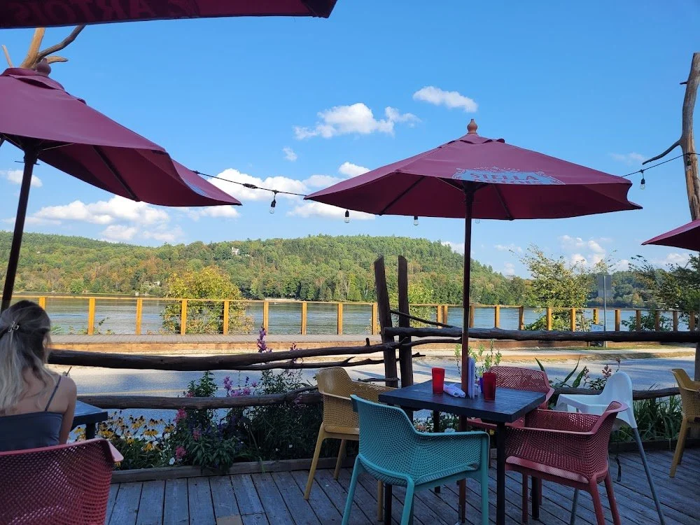 Outdoor patio with colorful chairs, red umbrellas, and a lake with trees and rolling hills in the background.