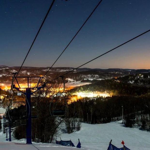 Nighttime view of a ski slope with a ski lift, snow-covered ground, and illuminated town lights in the distance under a clear, starry sky.