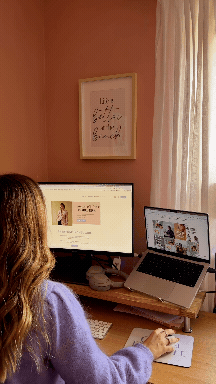 A person using a computer and laptop at a desk in a room with pink walls, a framed picture, and a window with white curtains.