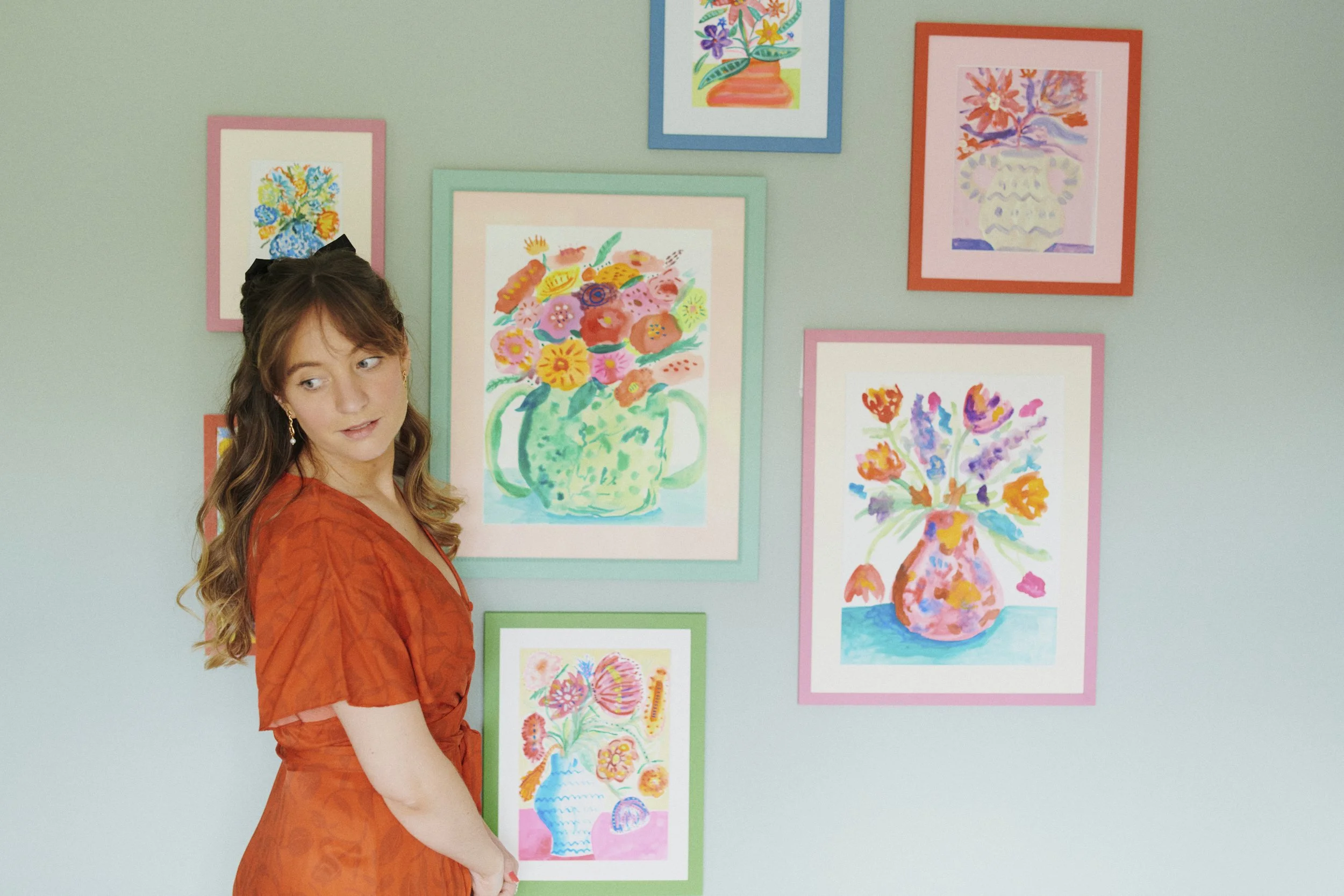 A woman in an orange dress standing in front of a wall decorated with colorful framed floral watercolor paintings.