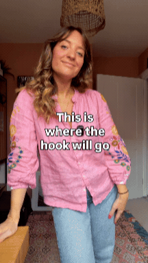 A woman with wavy blonde hair wearing a pink floral jacket and blue jeans, standing in a room with a white door and a patterned rug, smiling at the camera.