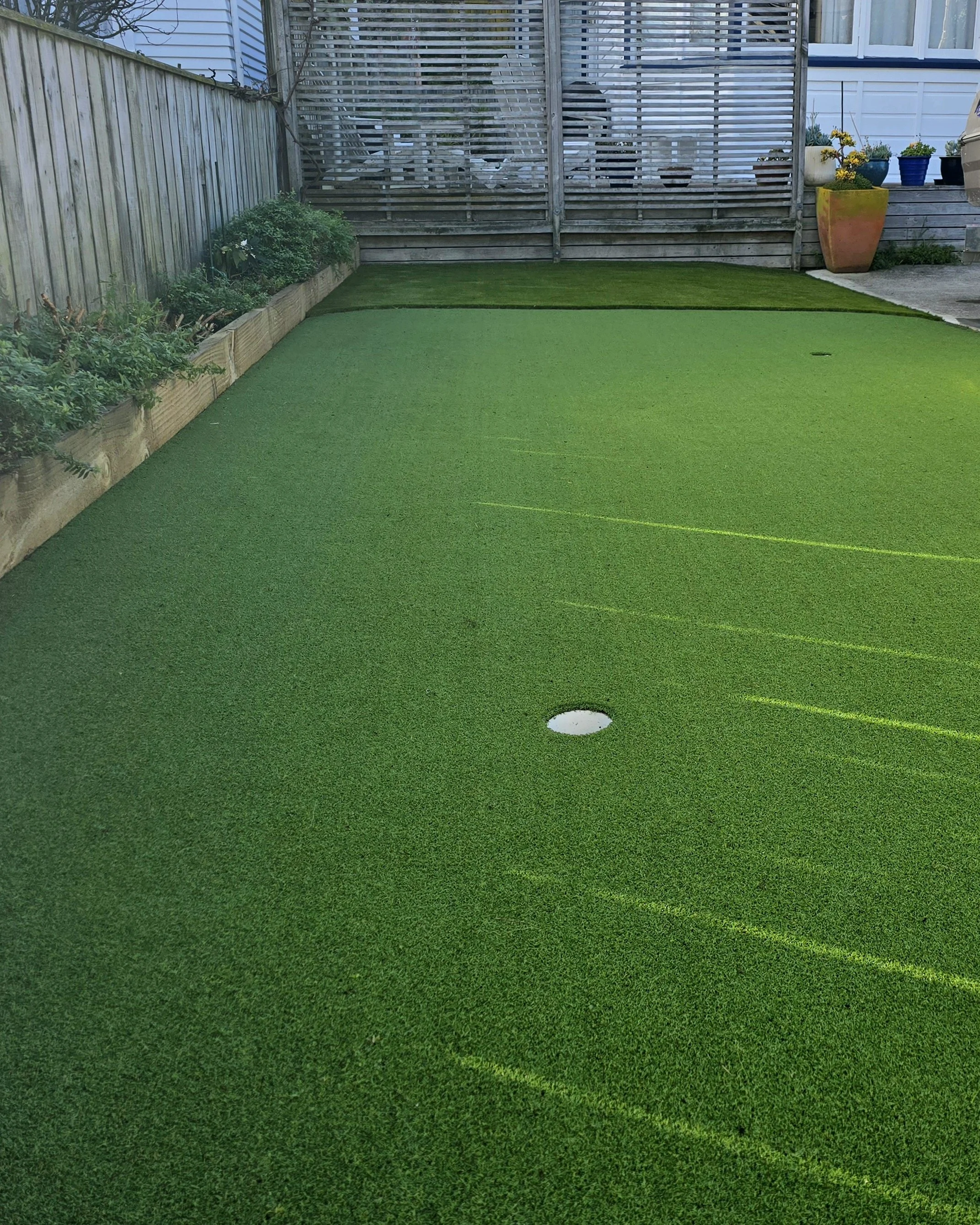 A backyard putting green with artificial grass, and a wooden fence with potted plants in the background.