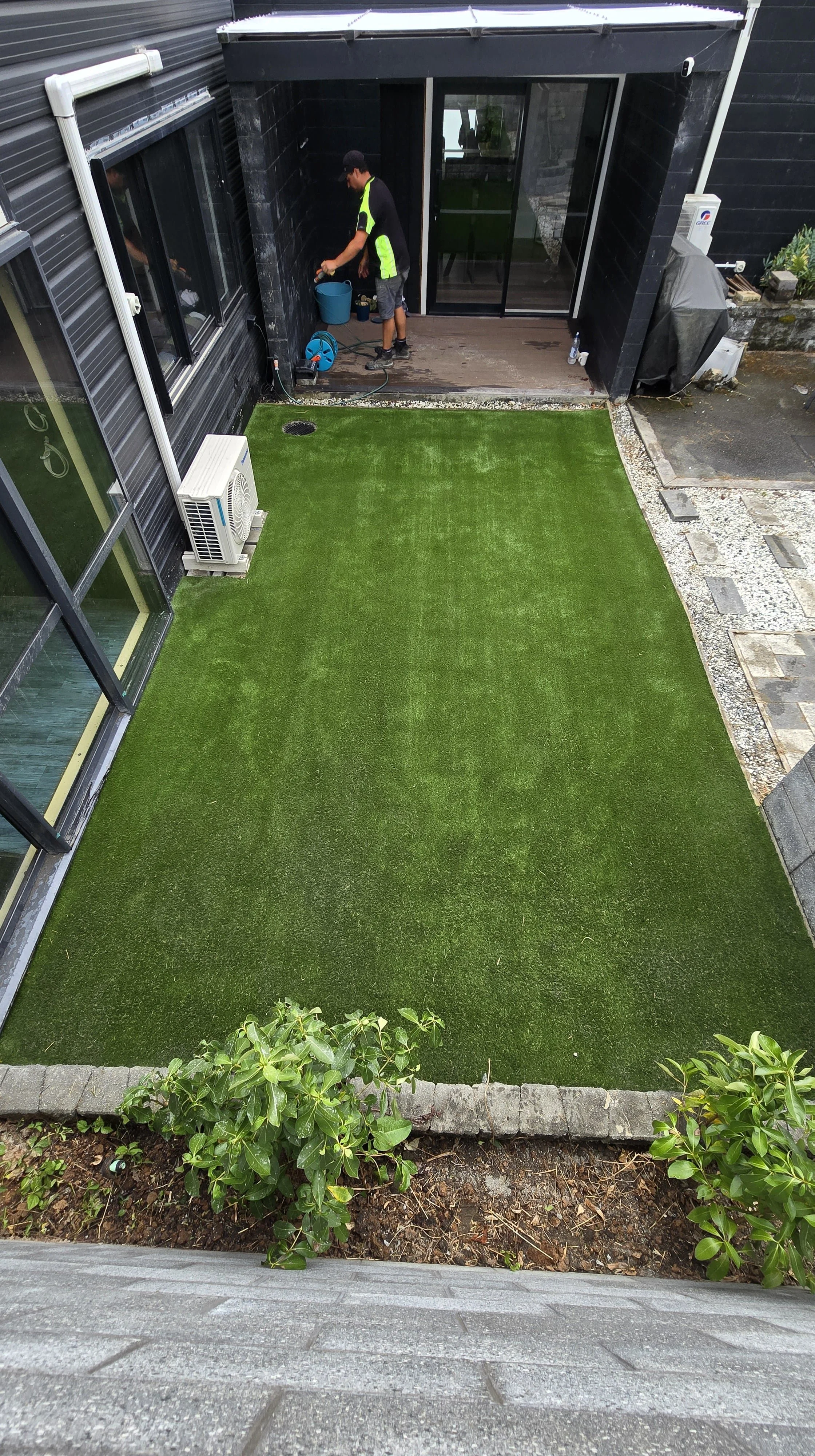 A person watering a small backyard with artificial grass, black exterior walls, and a sliding glass door. There is a window on the left with an air conditioning unit outside, and a small garden bed with plants at the bottom of the image.