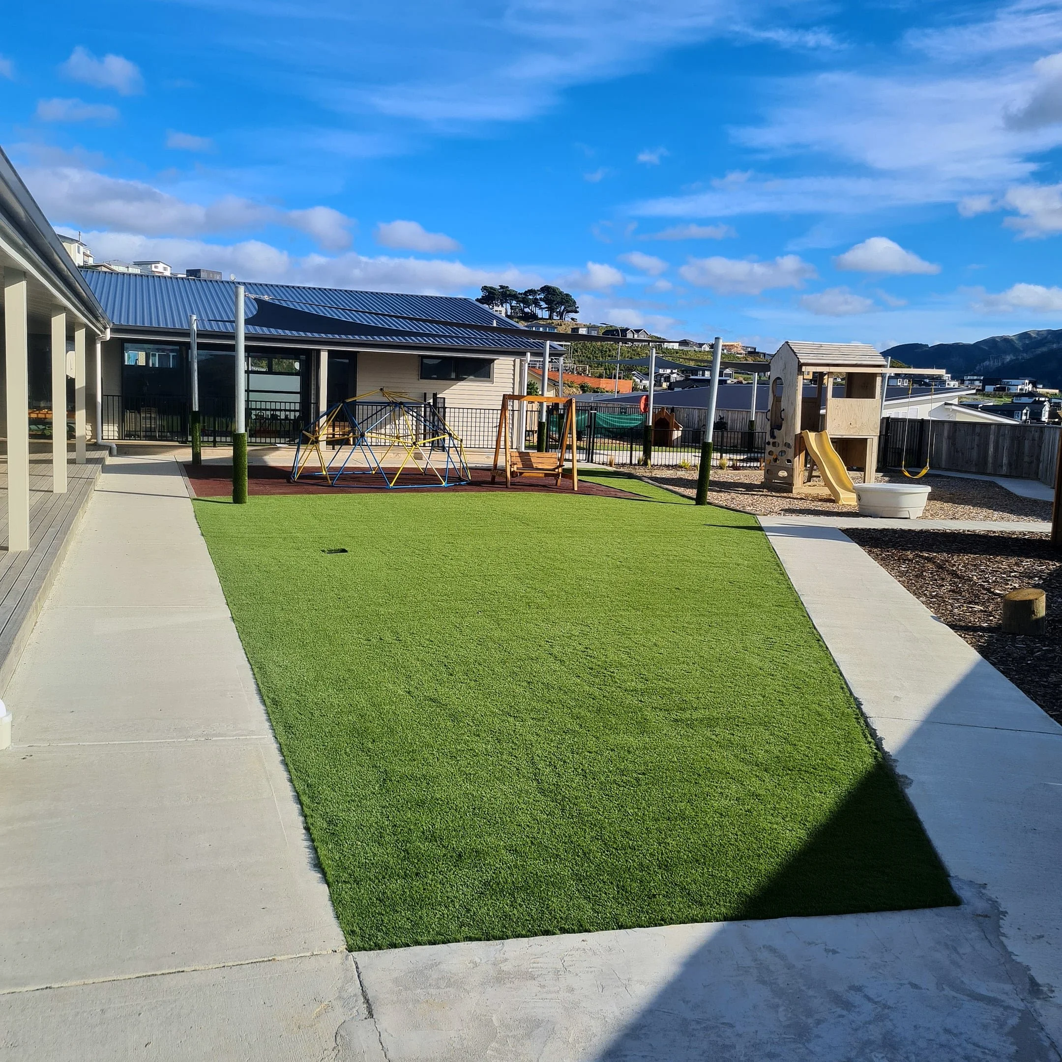 Childrens playground with artificial grass, wooden playhouse, slide, and climbing equipment, outdoor area in a residential neighborhood with houses and mountains in the background under a blue sky.