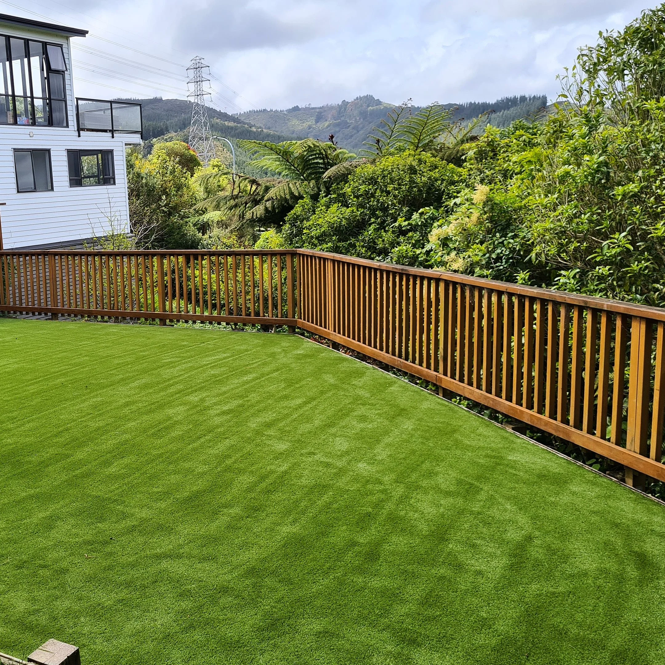 Green backyard with a artificial lawn and a wooden fence, surrounded by lush trees and a hillside in the background.