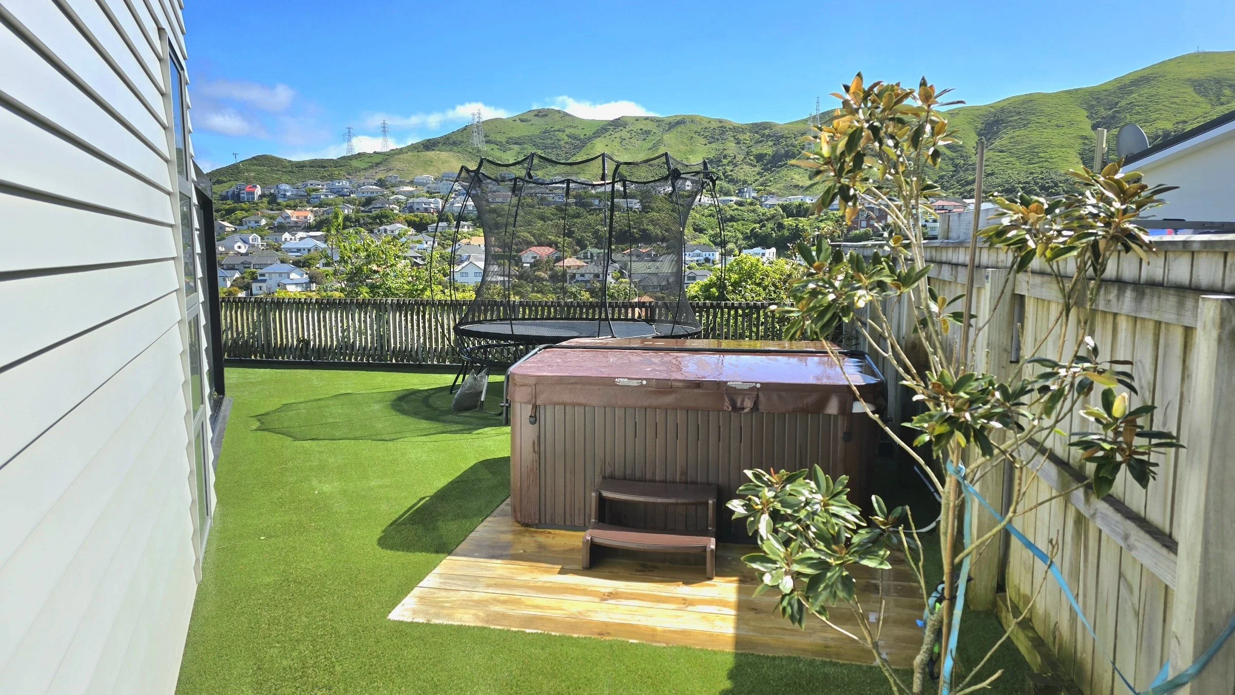 Backyard with hot tub on wooden deck, trampoline, small tree, artificial grass, overlooking hilly neighborhood under blue sky.