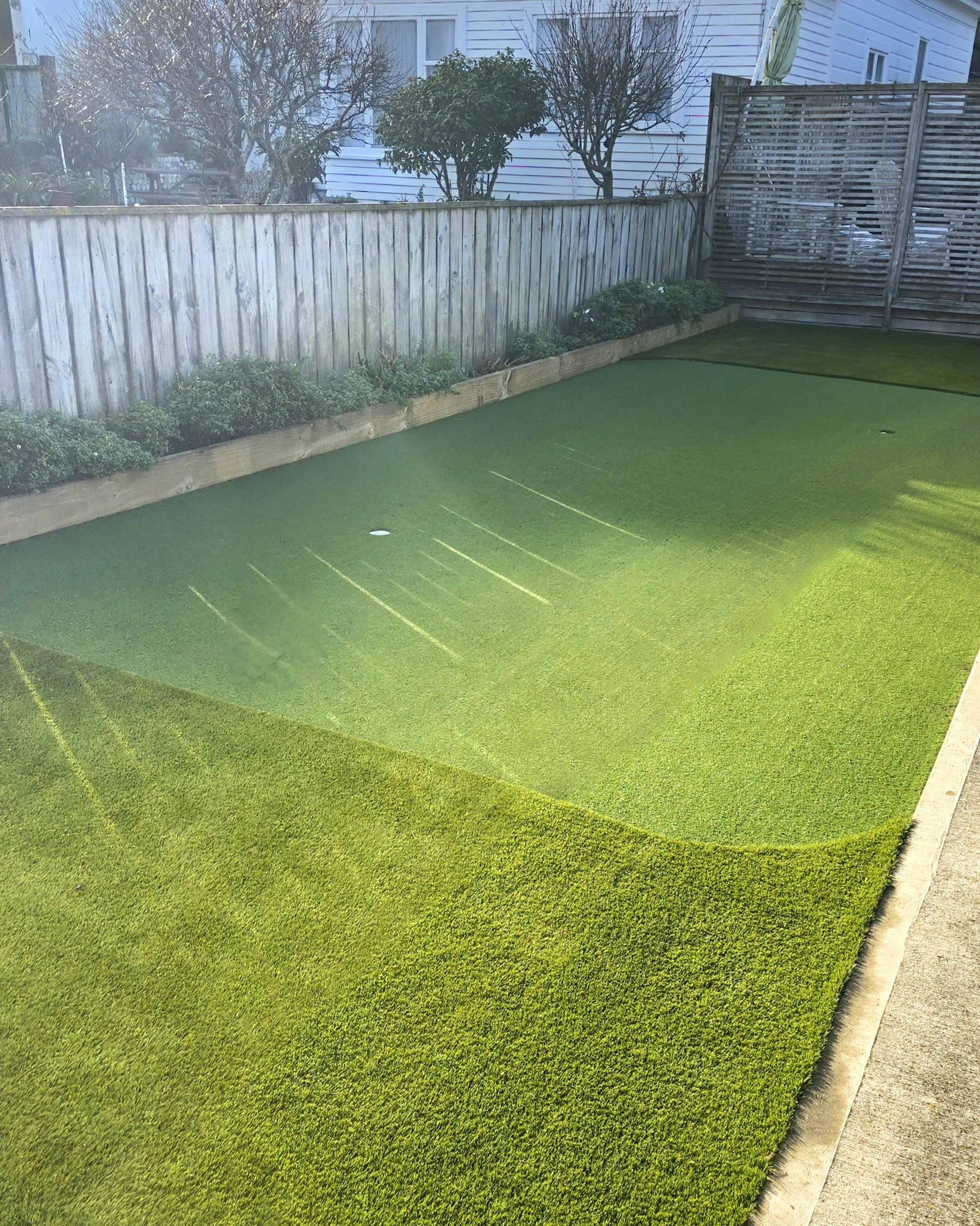 Backyard with artificial grass putting and chipping green, wooden fence, small bushes, trees, and a wooden privacy screen in the corner.