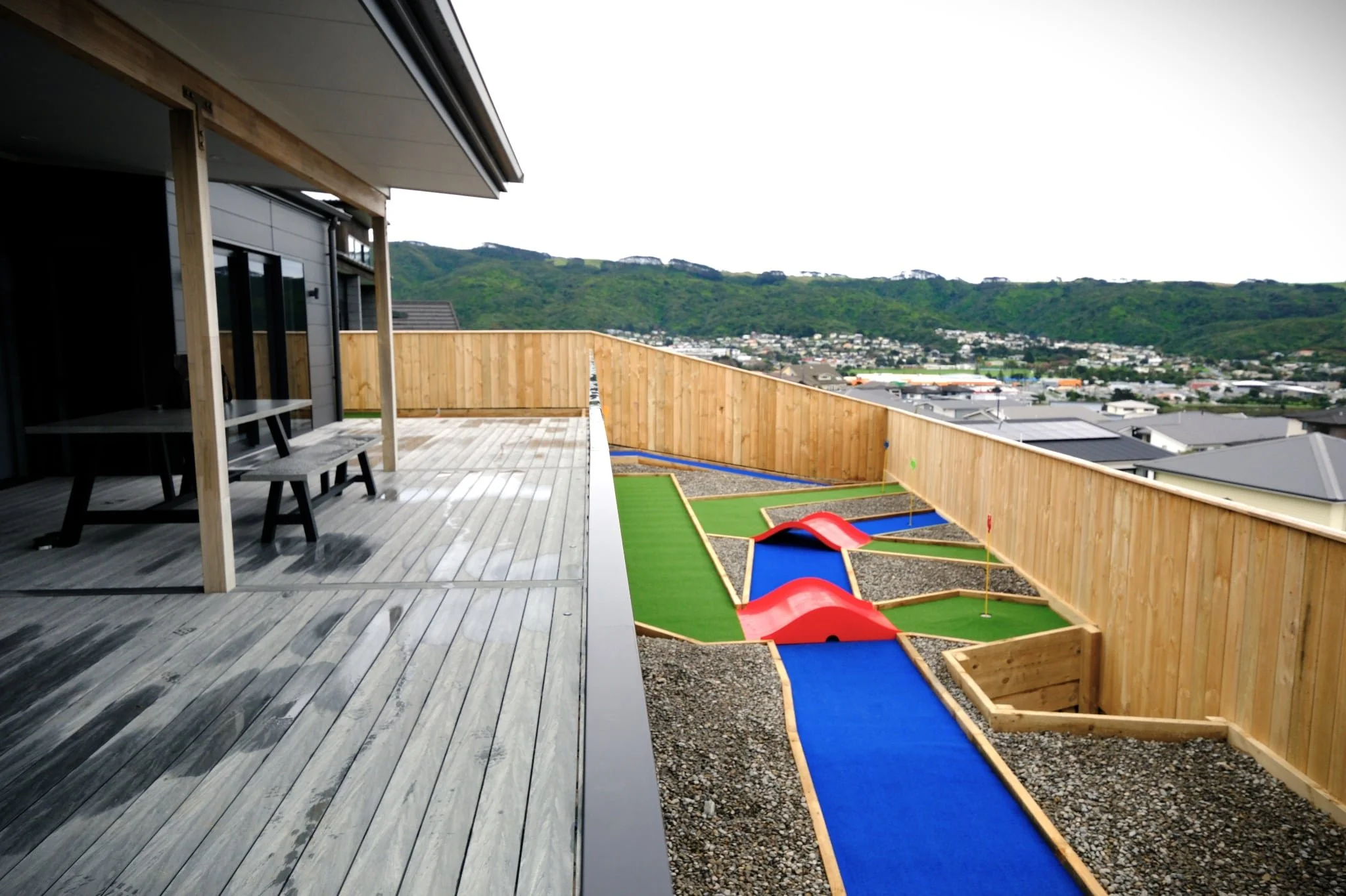 View of a rooftop patio with wooden flooring and outdoor furniture, overlooking a backyard minigolf course with red ramps and blue pathways, bordered by a wooden fence, with green hills and a town in the distance.